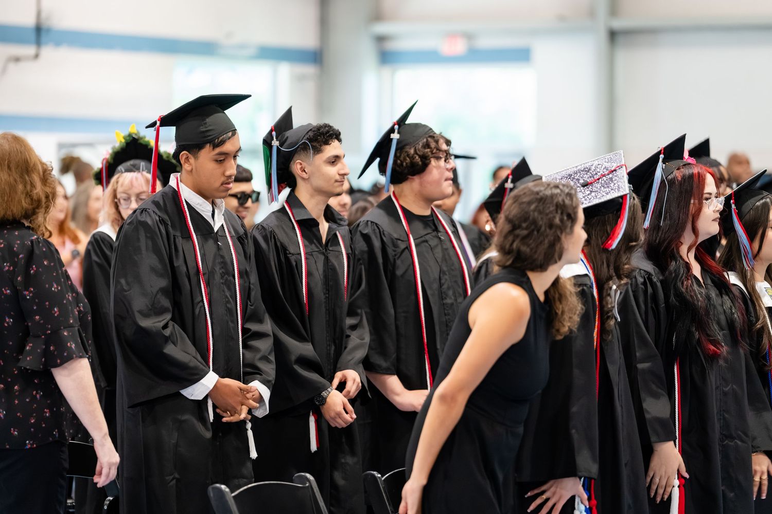 Students in black graduation gowns and caps stand in rows during a commencement ceremony indoors.