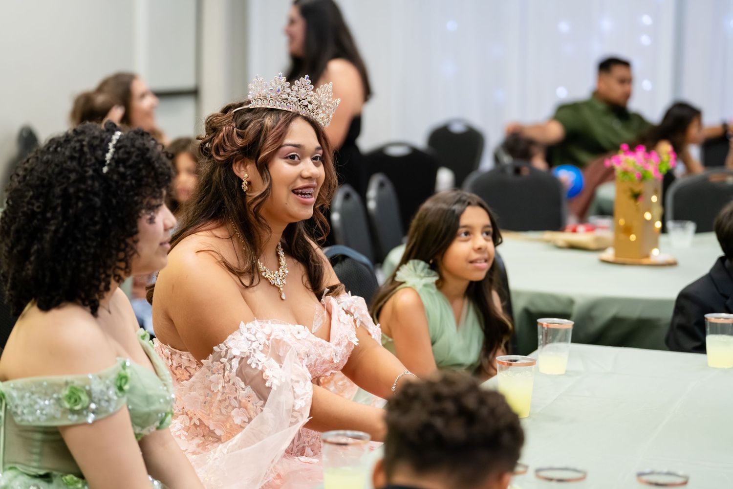 Young woman wearing a crown and pink gown smiles at a quinceañera celebration with guests seated at decorated tables.