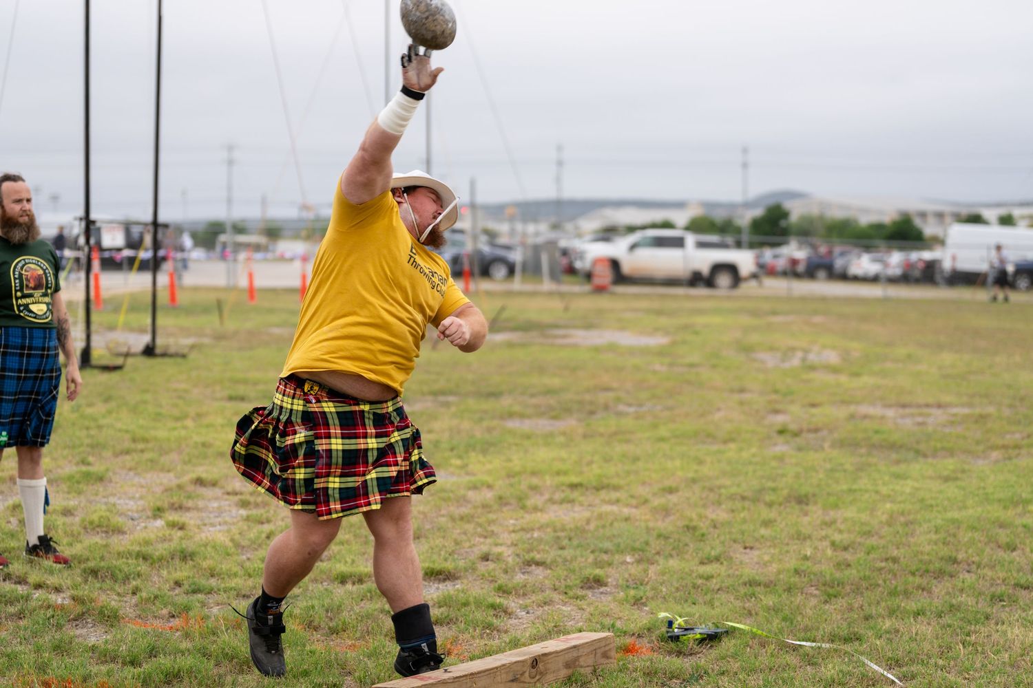 Man in yellow shirt and tartan kilt throws shot put at Highland Games competition outdoors.