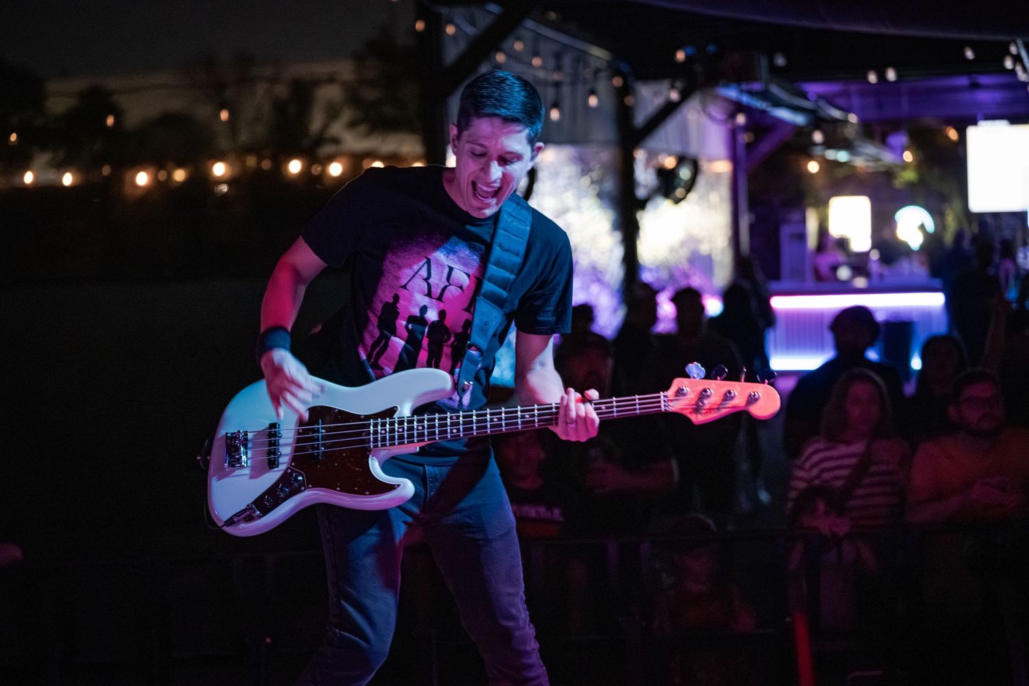 Musician playing white bass guitar on stage at night with colorful purple lights and crowd in background.