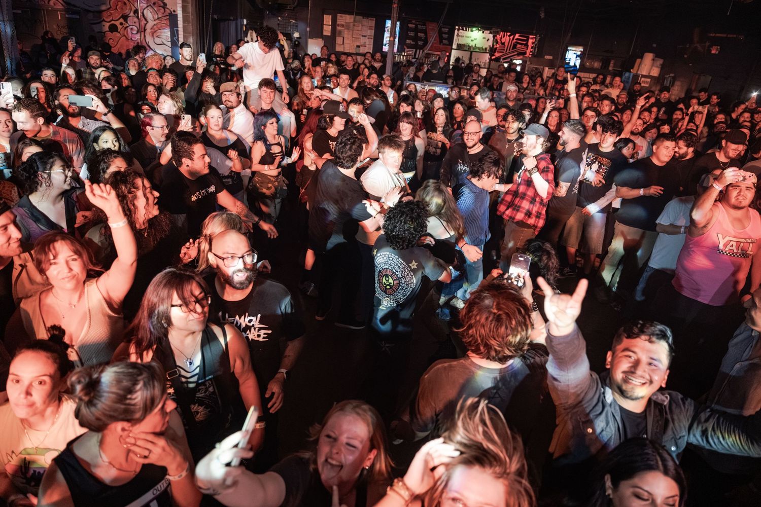Energetic crowd at a packed indoor rock concert, fans cheering and throwing up rock signs under warm venue lighting.
