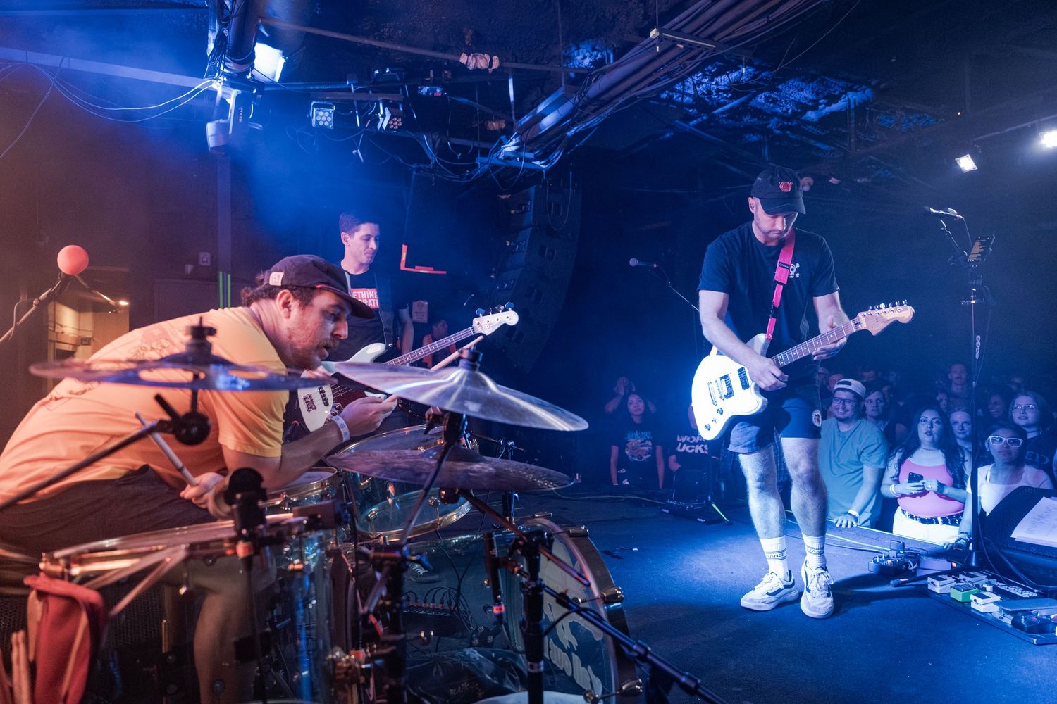 Band performs live on stage with drummer and guitarist under blue stage lights before an audience.