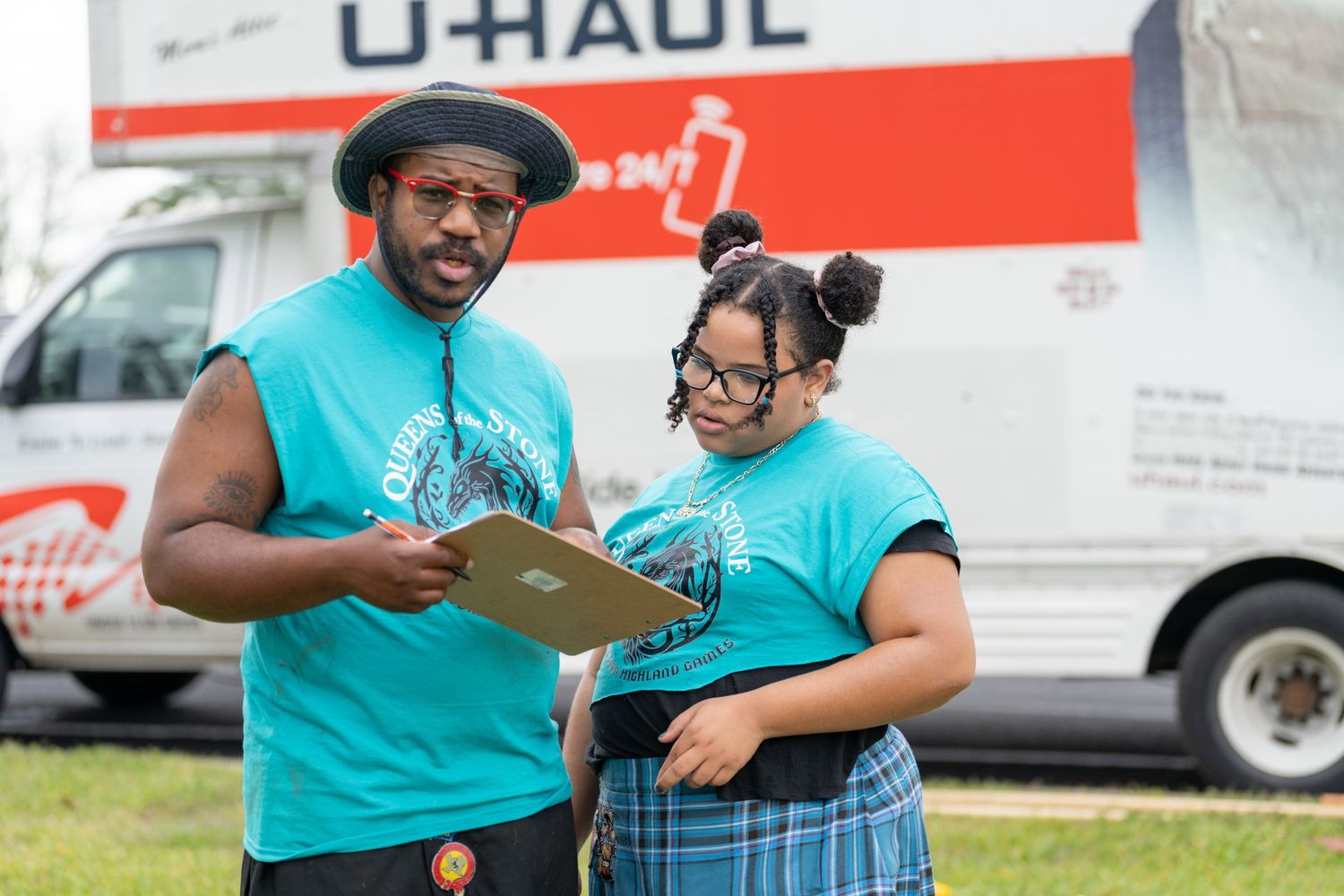 Two people in matching teal shirts review clipboard near U-Haul moving truck outdoors.