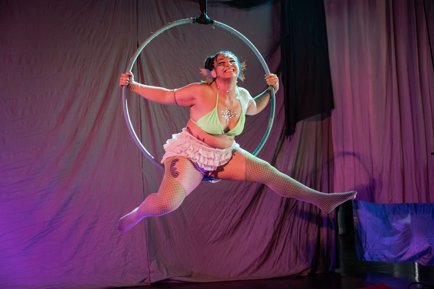 Aerial hoop performer in white costume striking a dynamic pose on a lyra ring under colorful stage lighting.