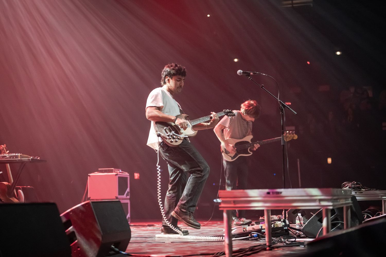 Two guitarists perform energetically on a dramatic red-lit concert stage with spotlights beaming overhead.