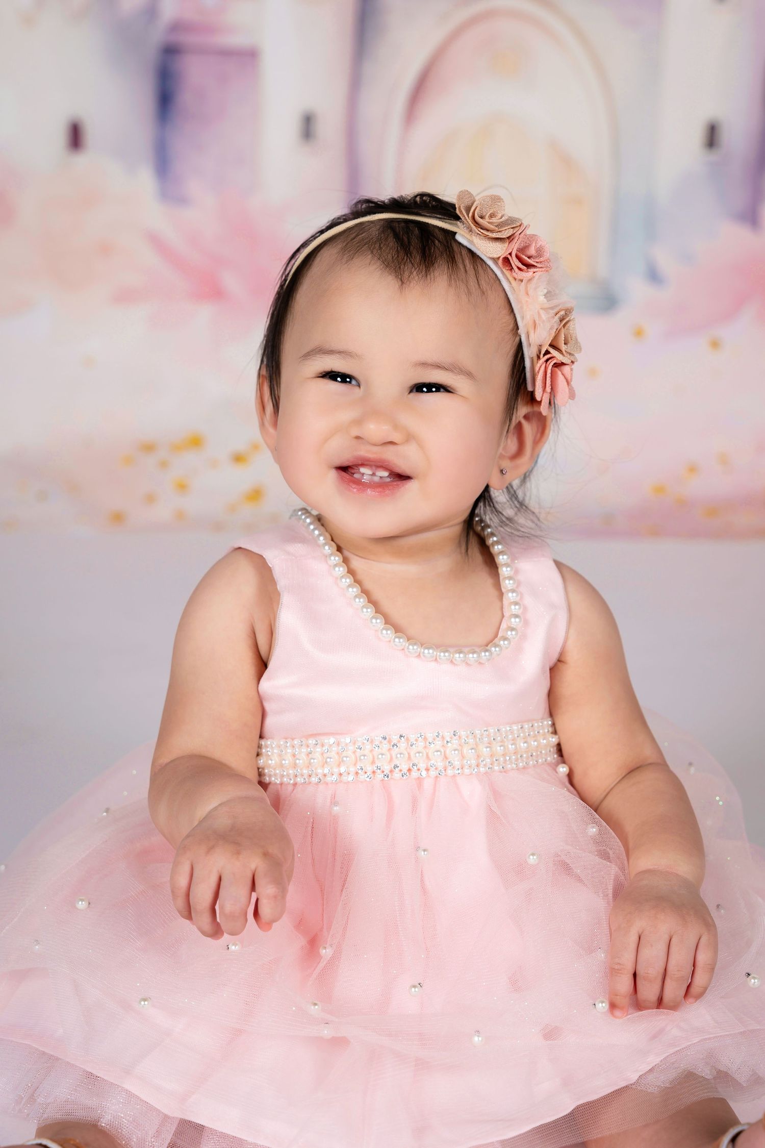 Smiling baby girl in pink tulle dress with pearl necklace and floral headband, posed in front of a pastel princess backdrop.