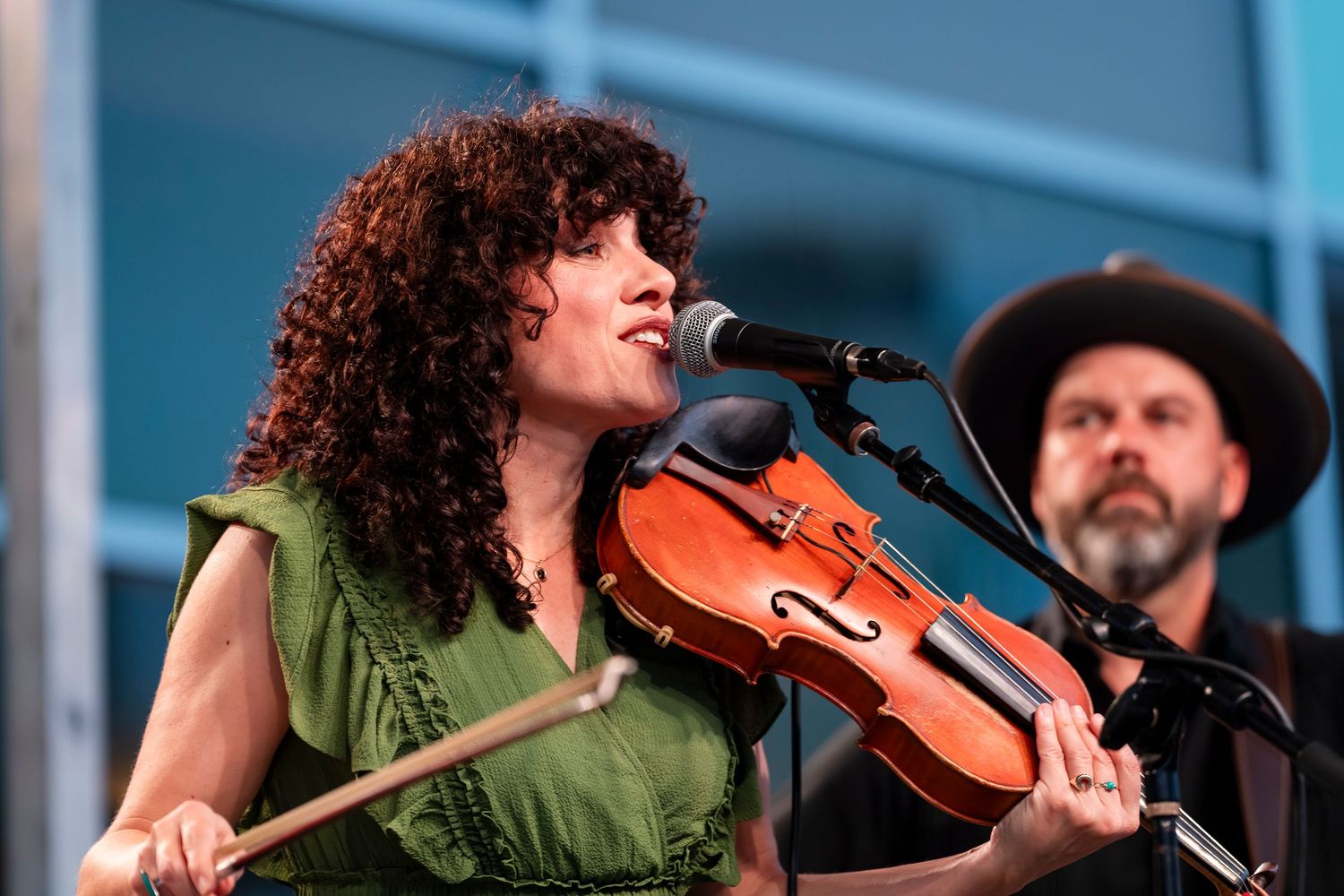 A musician with curly hair performs with a violin on stage in a green top.