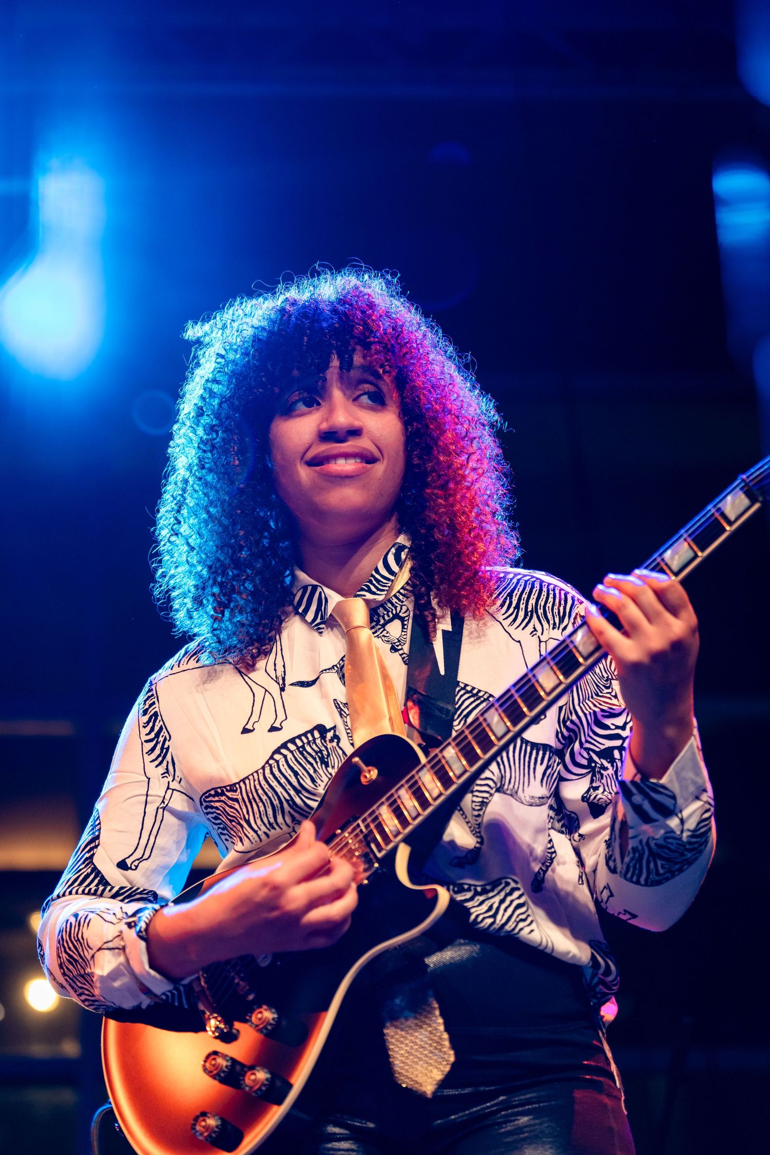 Musician with curly hair performs energetically on electric guitar under blue stage lighting.
