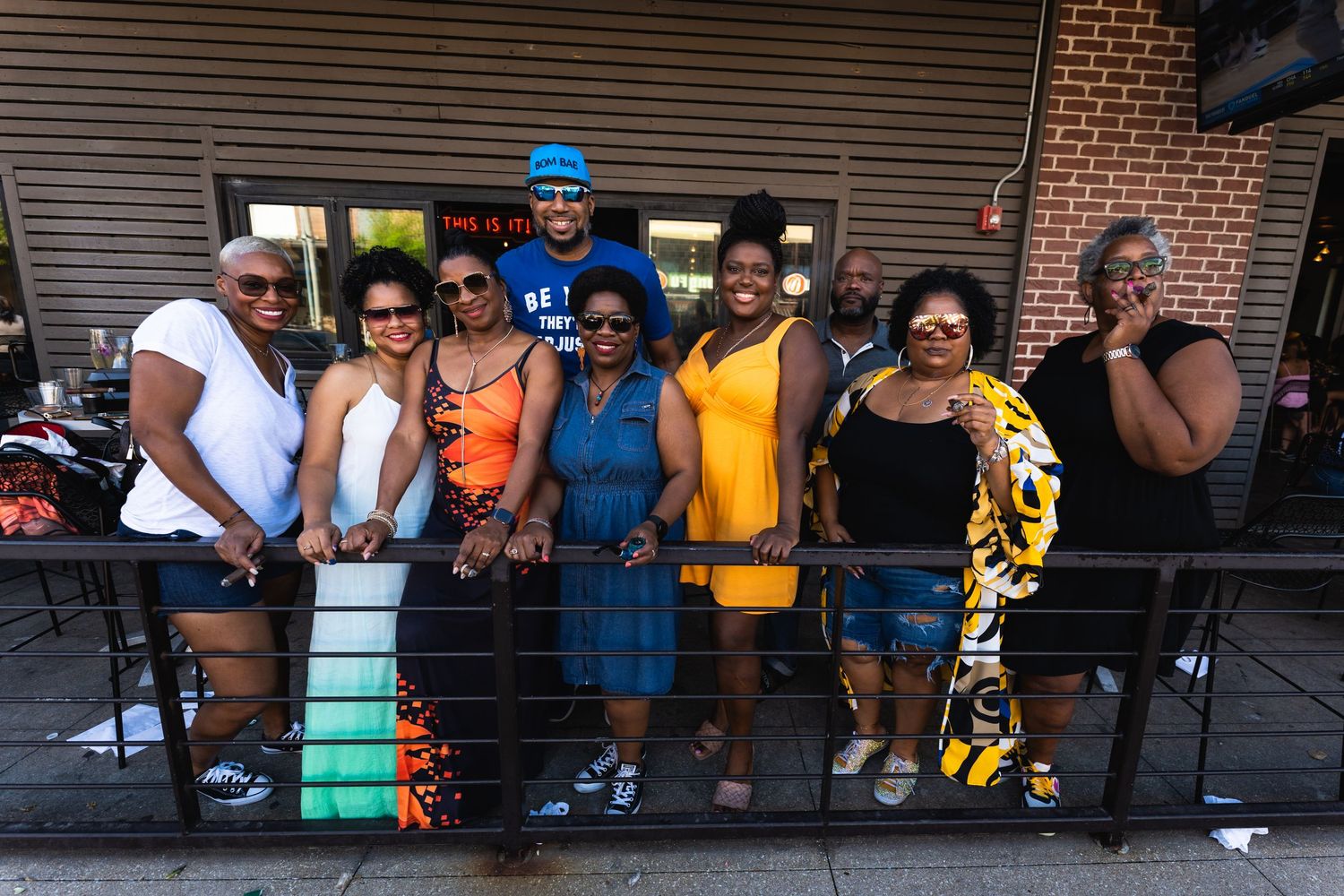 A group of friends in colorful summer outfits pose together on a patio railing outside a brick building.