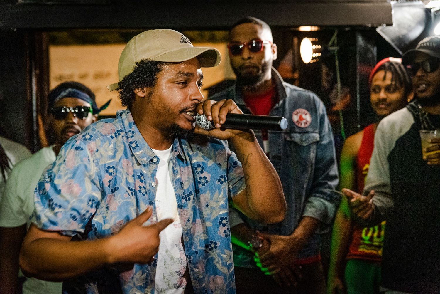 A performer in a patterned blue shirt and baseball cap holds a microphone while entertaining a crowd at an indoor venue.