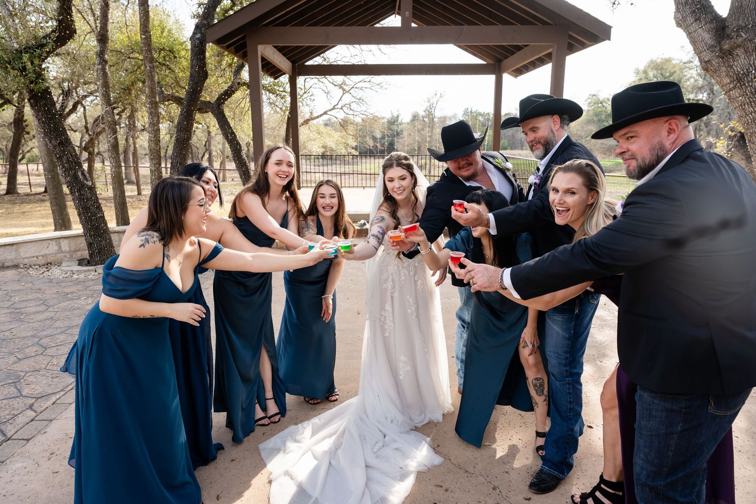 Wedding party toasting outdoors under a wooden gazebo, bridesmaids in navy dresses and groomsmen in black cowboy hats.