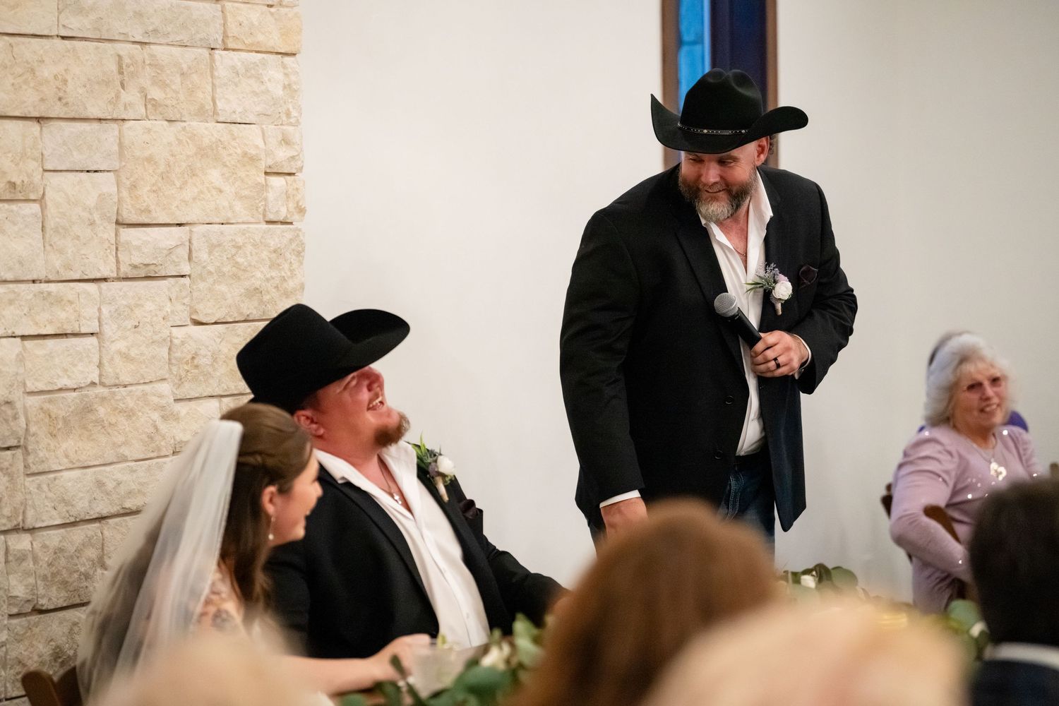 Man in black cowboy hat and blazer speaks at a country wedding ceremony with guests in cowboy hats.