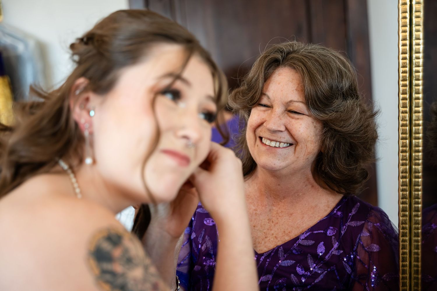 Bride putting on earrings while smiling woman in purple dress laughs beside her in a gold-framed mirror.