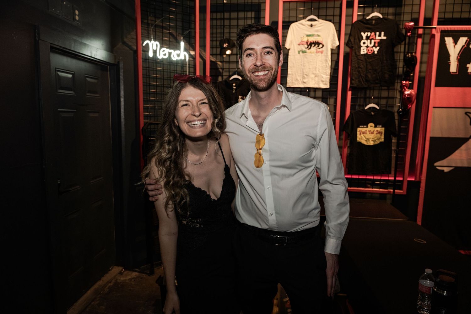 A smiling couple poses together at a dimly lit bar or restaurant with neon signs and merchandise displays in the background.