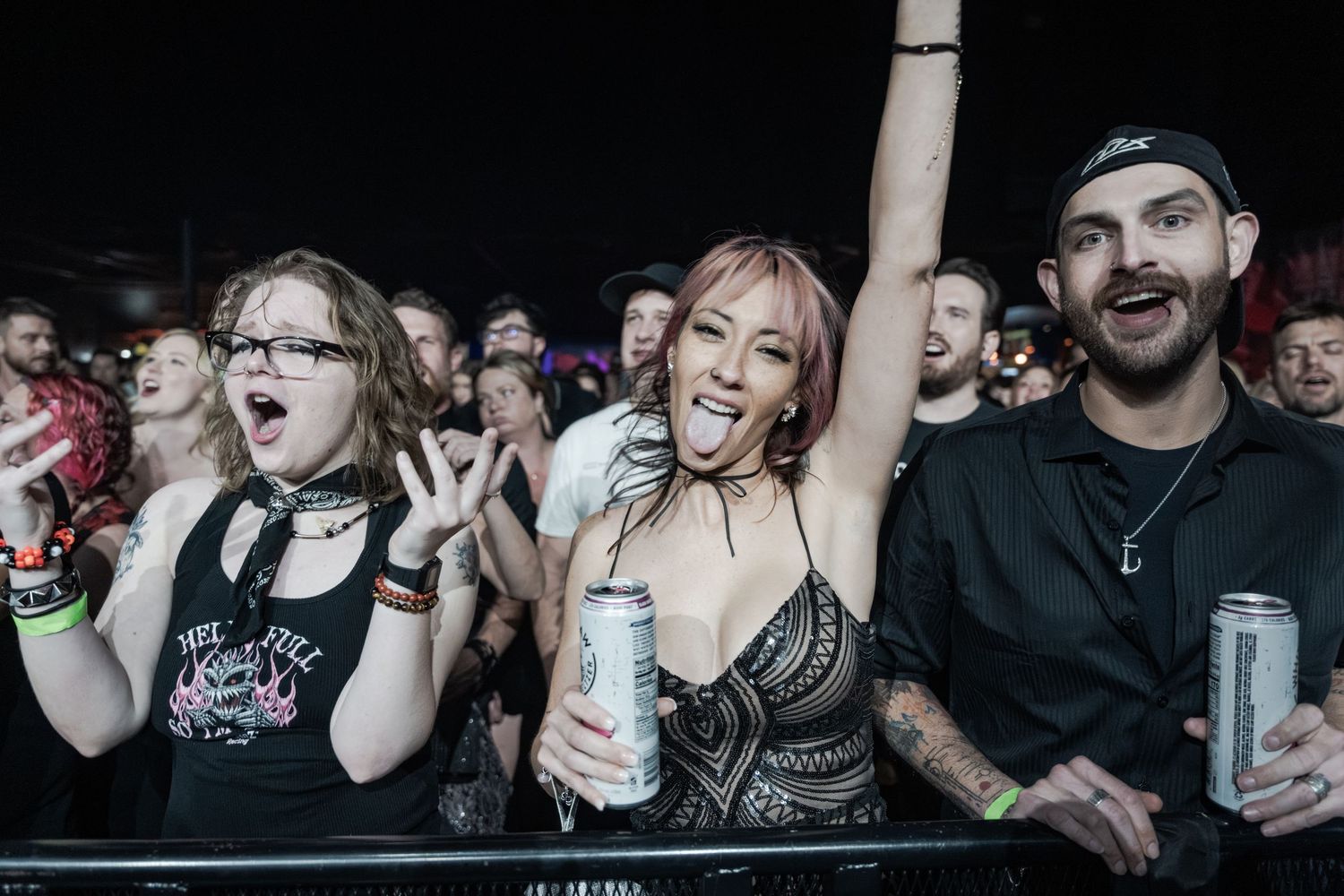 Excited concert crowd cheering at a live music event, woman raising arms holding beer can at dark venue.