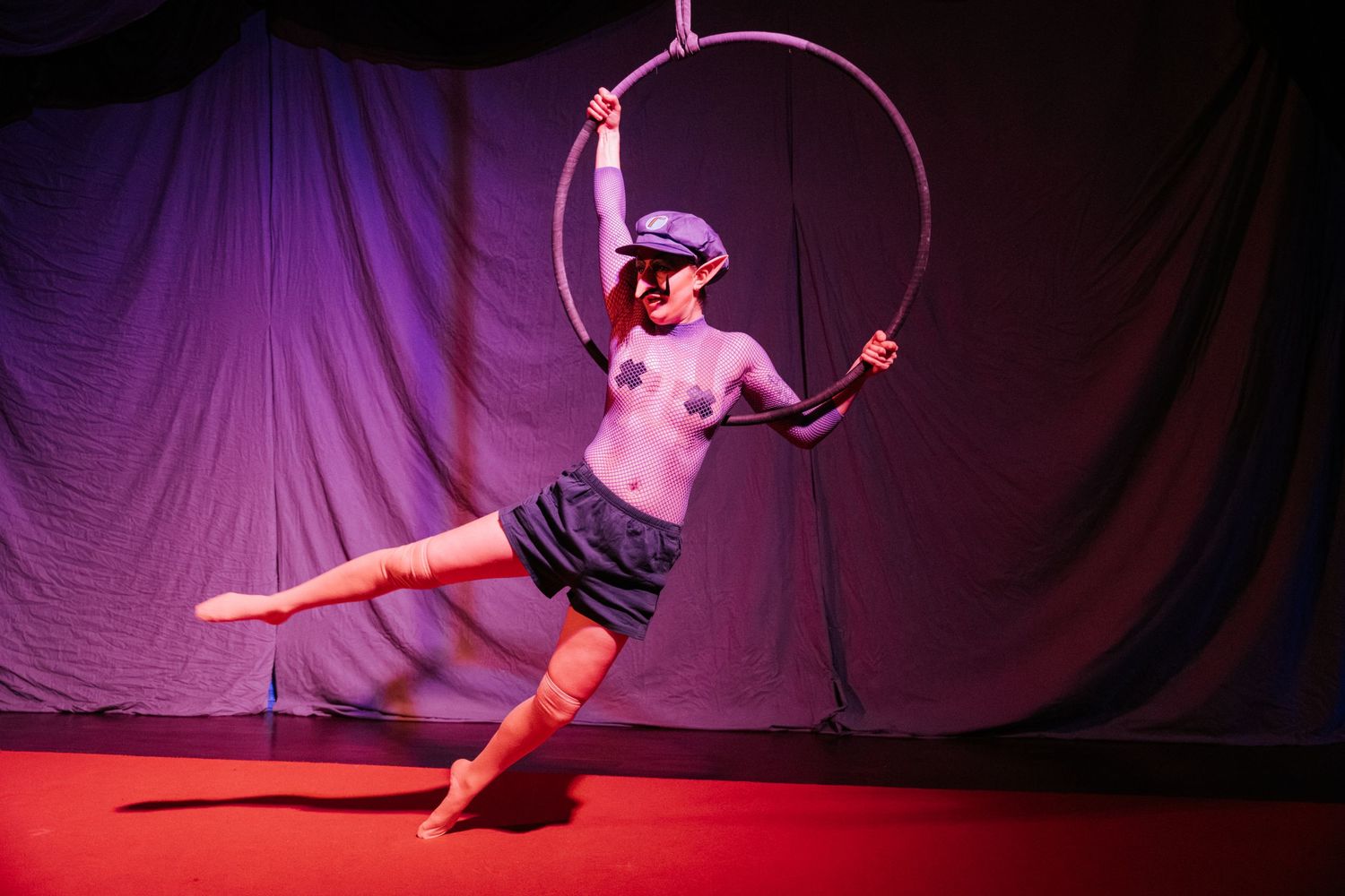 Aerial hoop performer in cap and shorts executing a graceful pose on a red-lit stage with purple curtains.