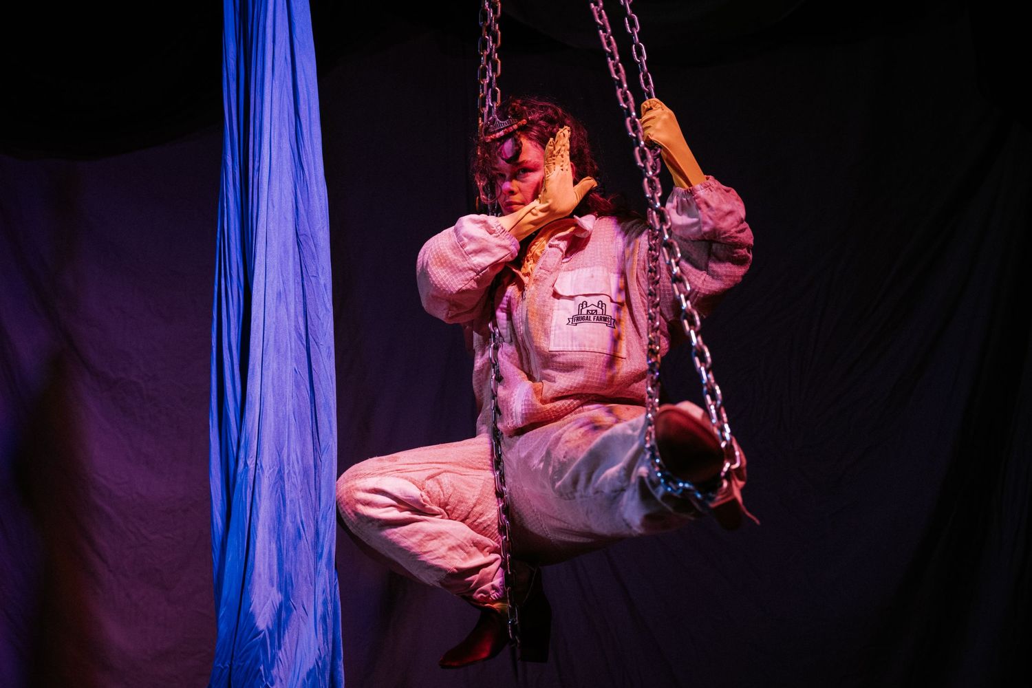 Performer in pink jumpsuit suspended on chains with blue silk fabric in dramatic stage lighting.