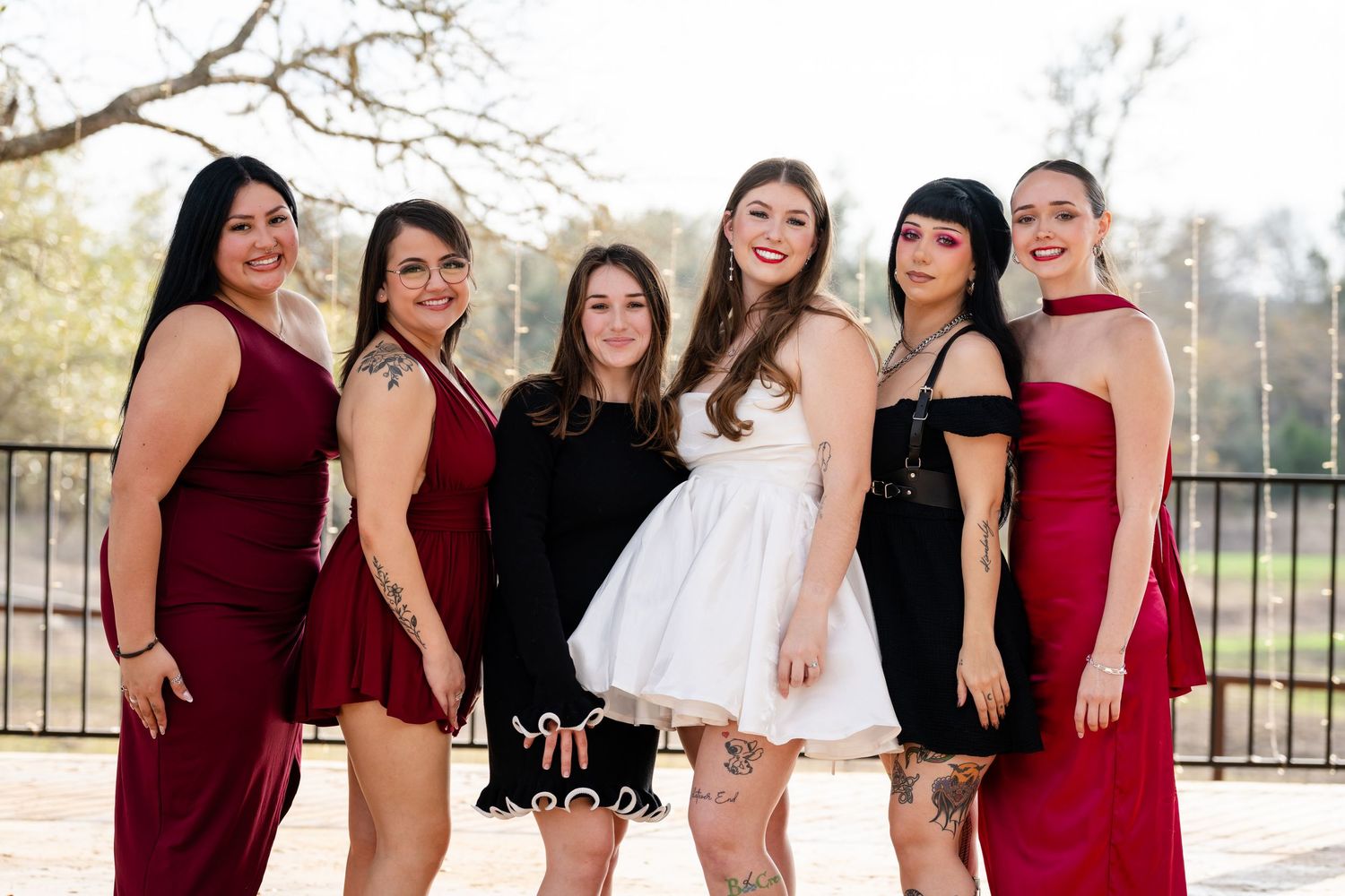 Six women in formal dresses posing together outdoors, wearing burgundy and black attire at a special occasion.