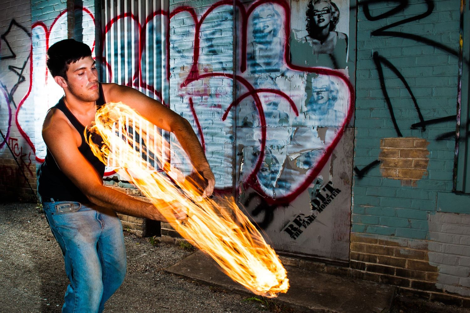 A person performs fire spinning tricks against a graffiti wall creating dramatic light trails in the dark.