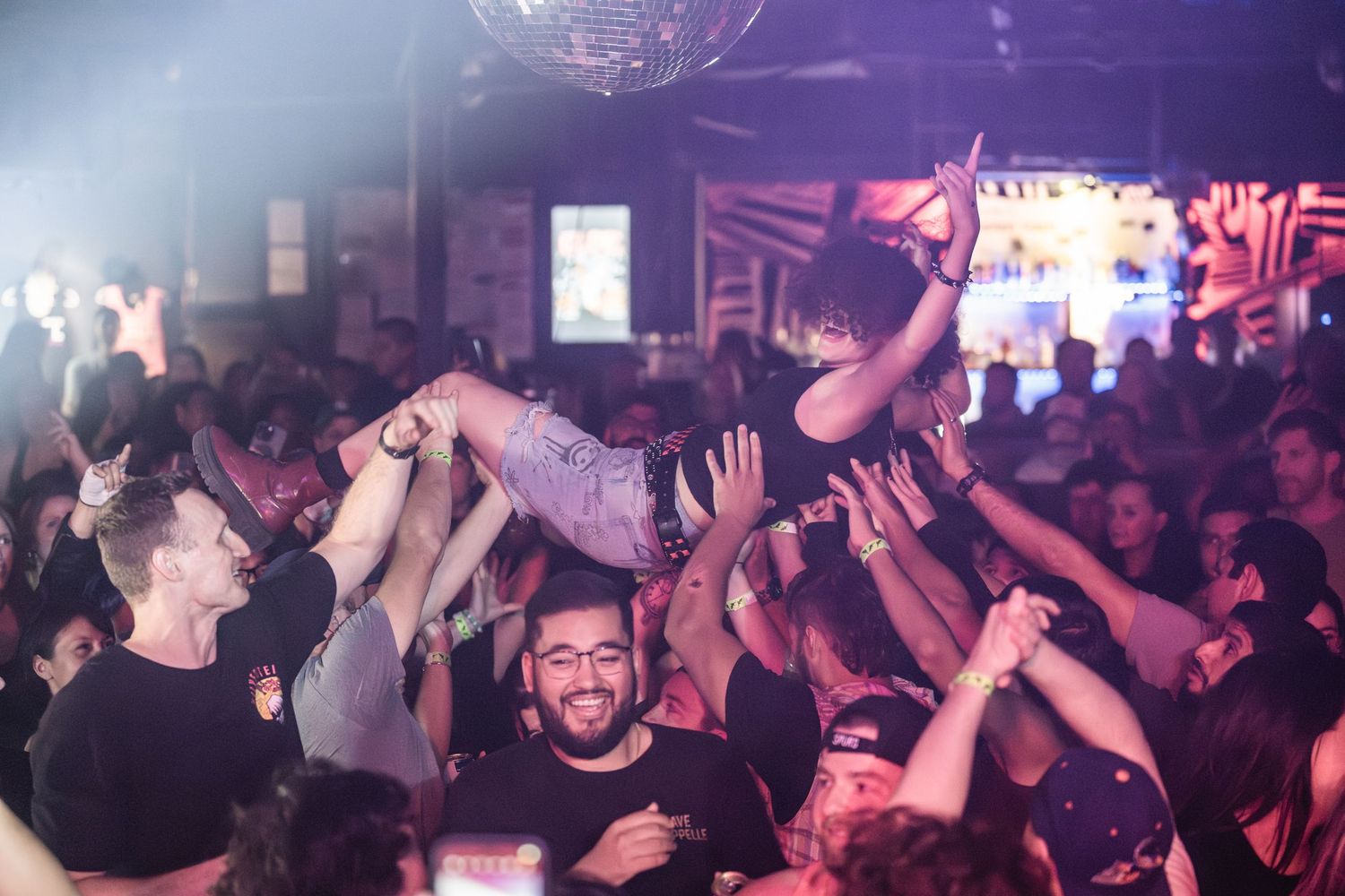 Crowded nightclub scene with purple lighting and people dancing with raised hands.