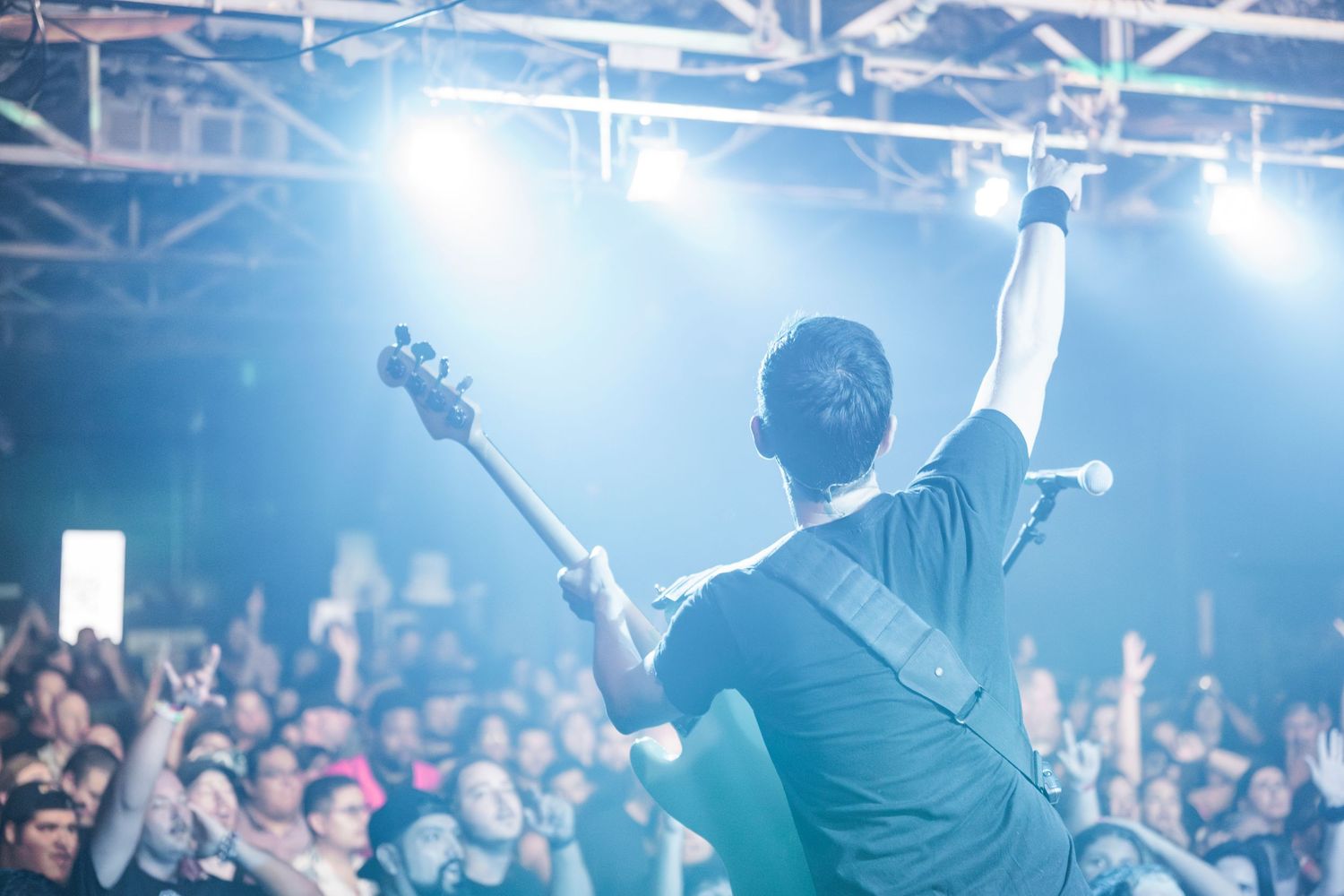 Silhouetted musician performs on stage with bass guitar in front of large crowd under blue stage lighting.