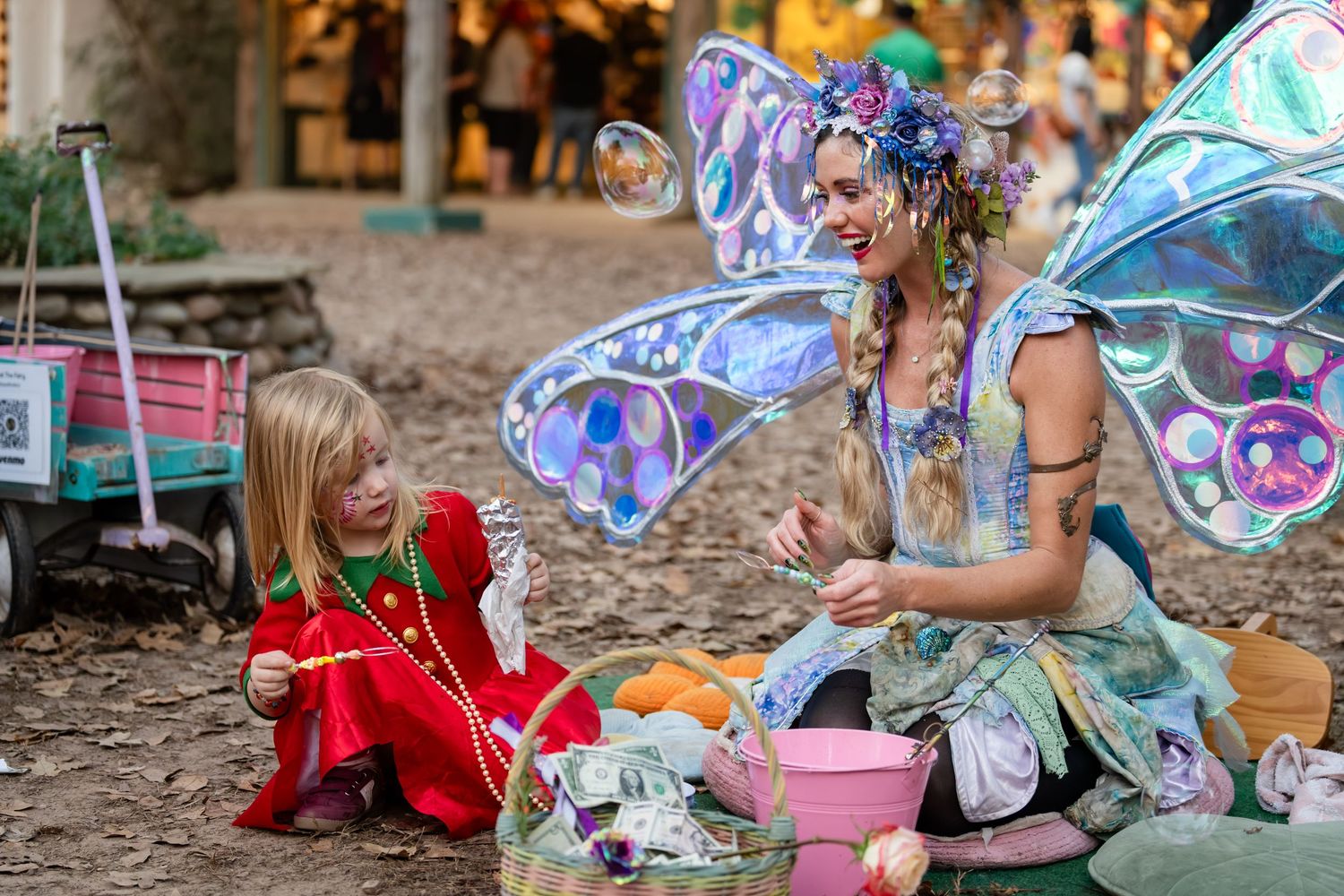 A magical fairy with iridescent wings interacts with a young child in red during an enchanting outdoor moment.