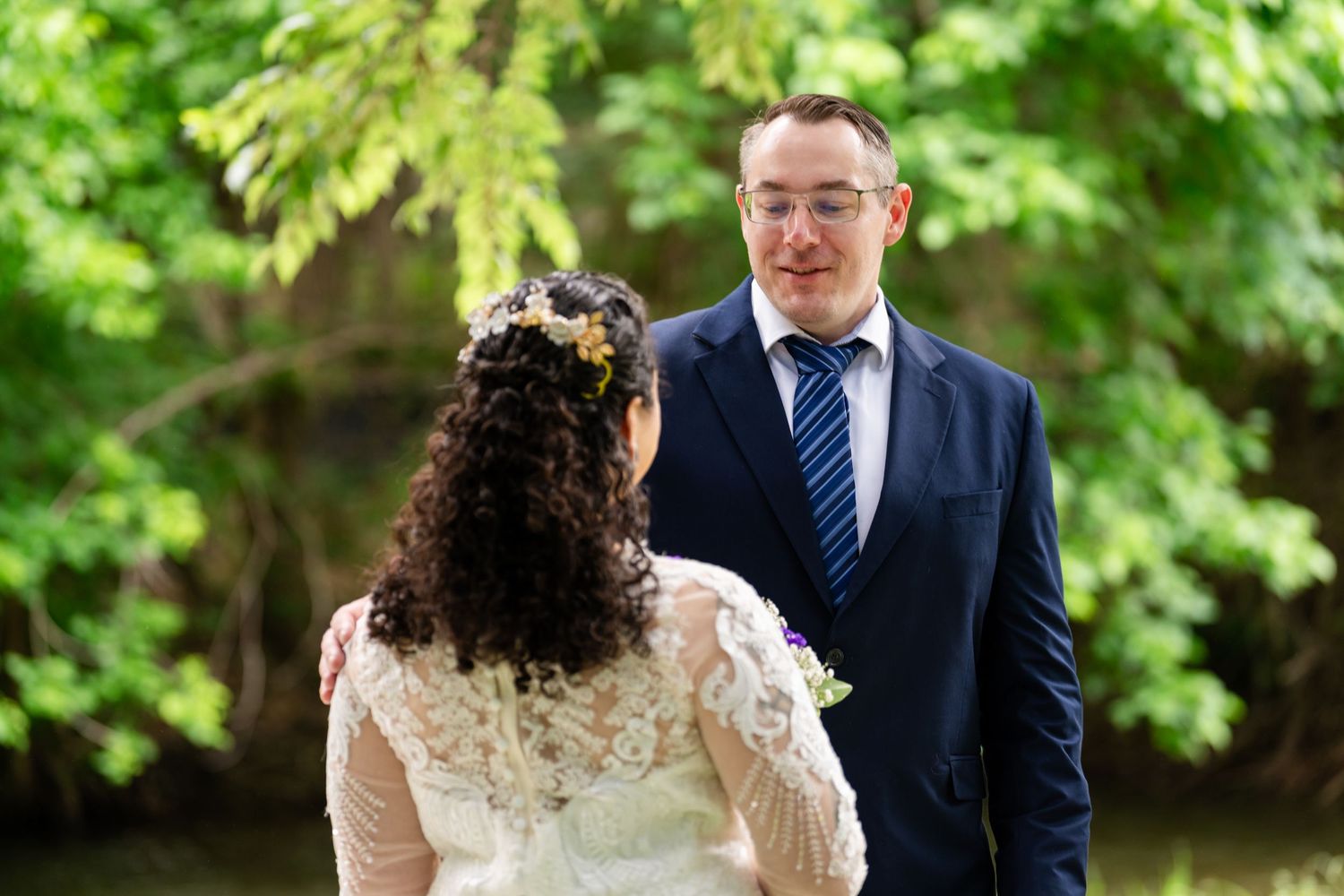 A bride and groom share a tender moment outdoors, surrounded by lush green trees, as the groom gazes lovingly at his bride.