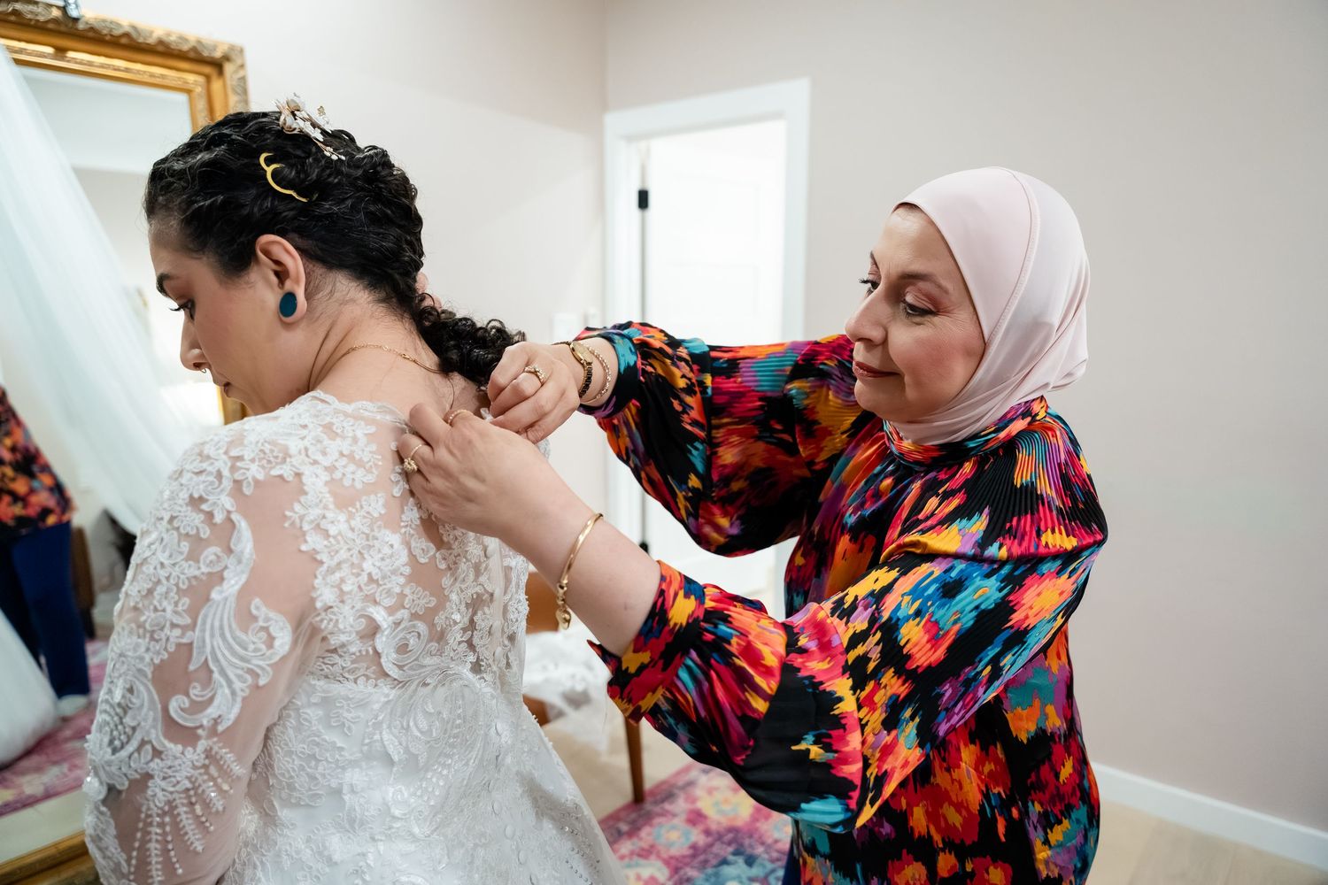 A woman in a hijab helps a bride in a lace wedding dress with her necklace in a bright, elegant room.