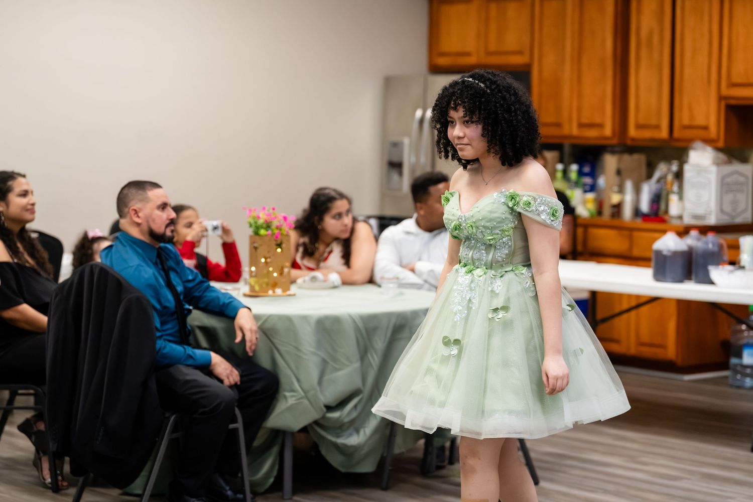 Young woman in elegant green dress walks at indoor fashion show with seated audience in background.