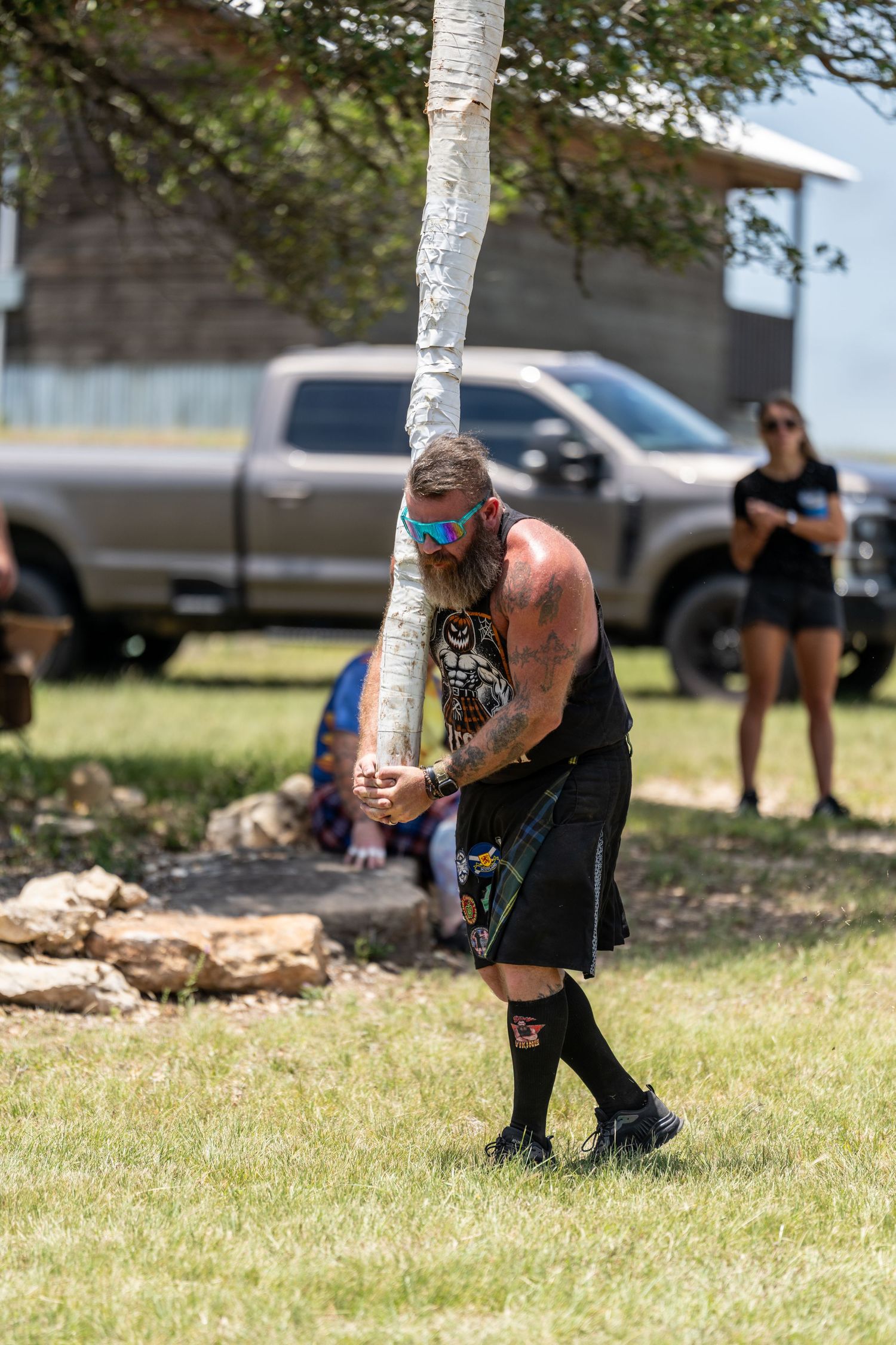 Tattooed man in kilt and knee socks tosses caber at Highland Games outdoor competition on sunny day.