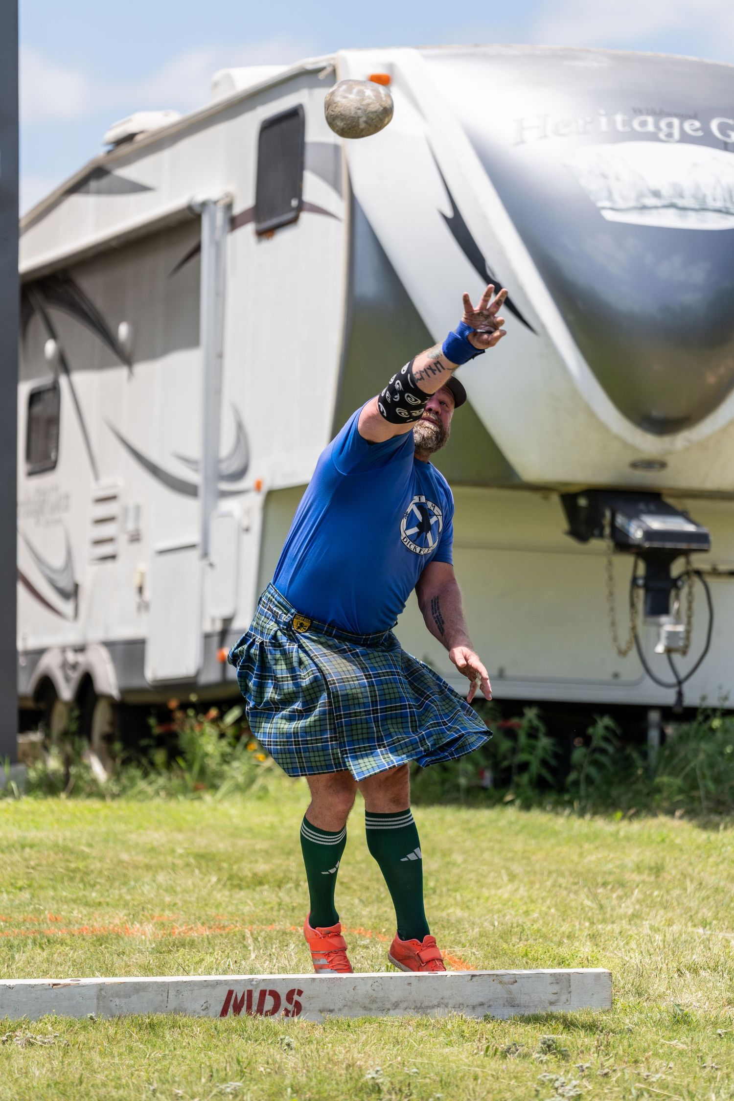 Man in blue shirt and tartan kilt throwing a stone ball at a Highland Games competition outdoors.