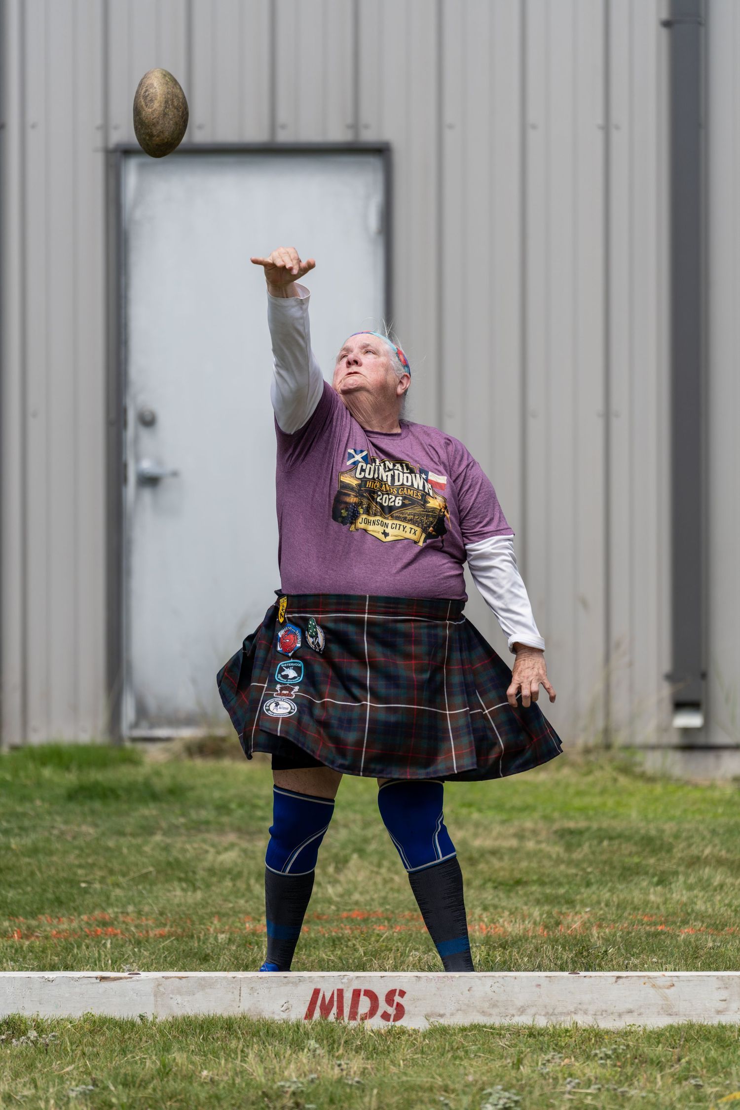Man in tartan kilt throwing a ball at a Highland Games competition outdoors.
