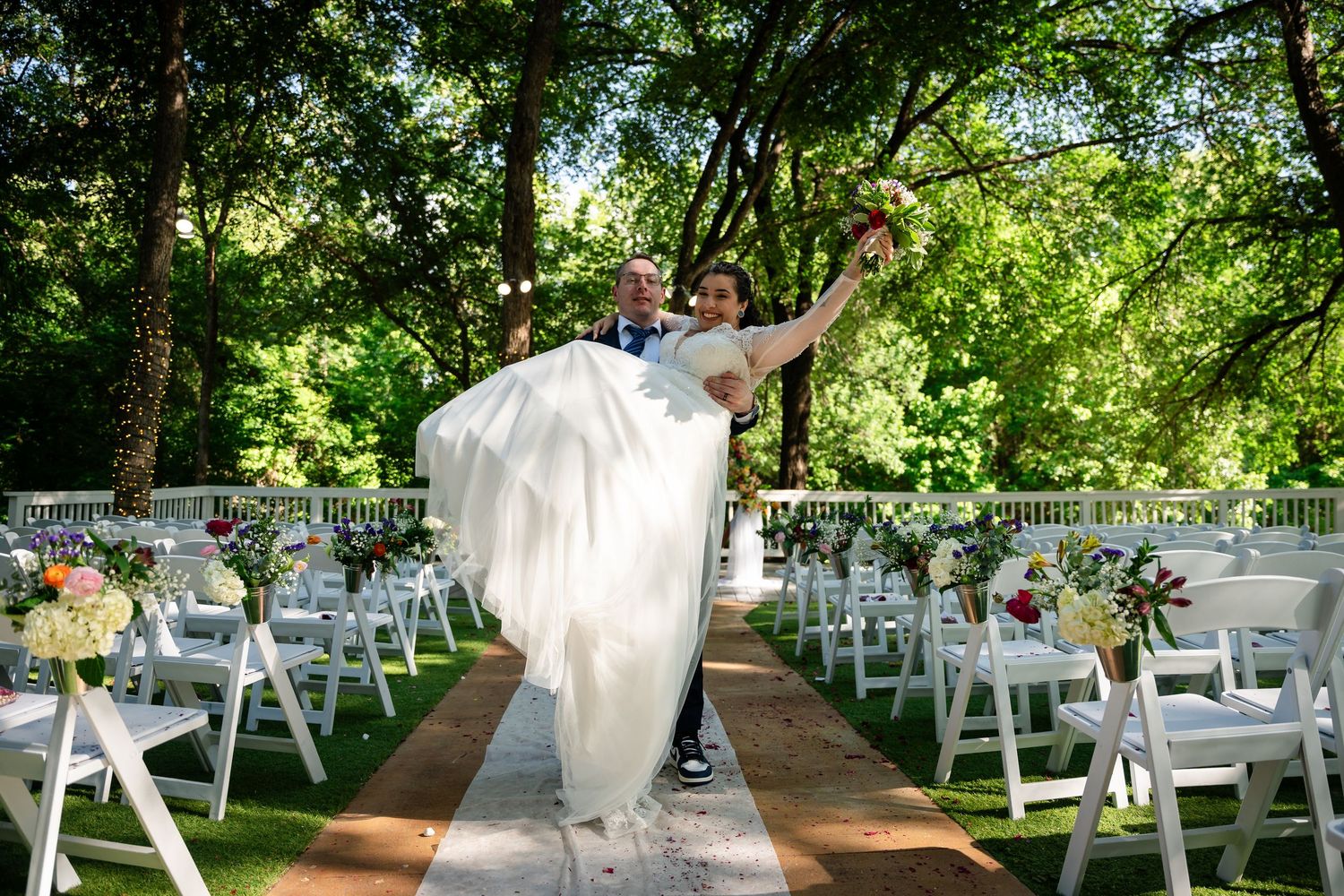 Groom lifting bride in white gown at outdoor wedding ceremony with white chairs and lush green trees.