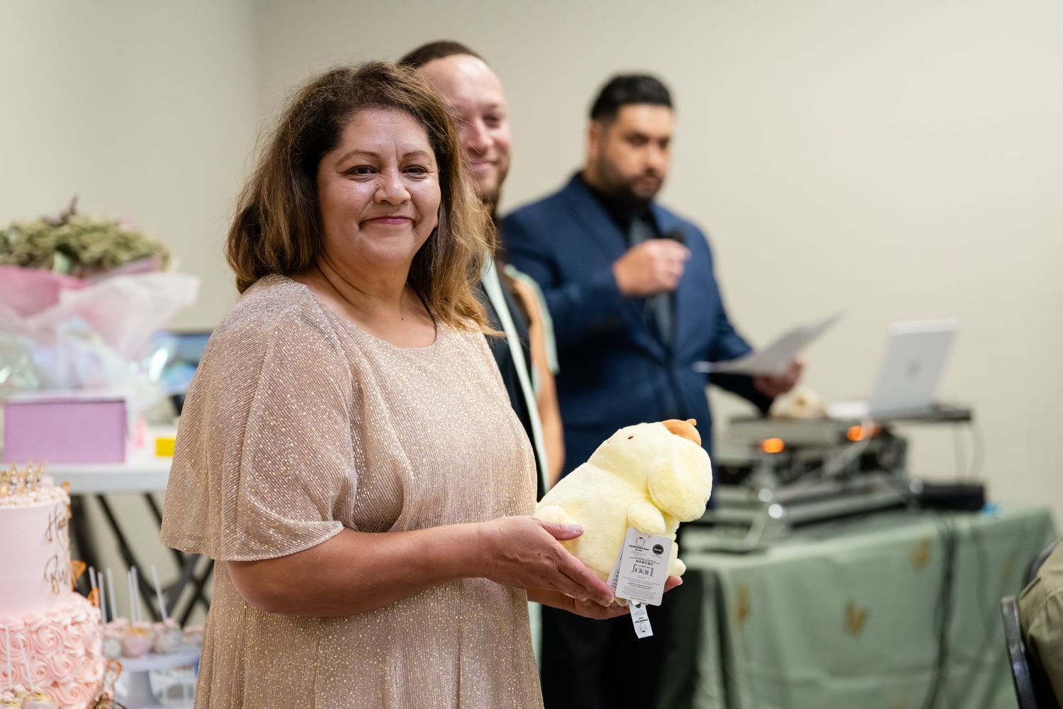 Woman smiling and holding a yellow gift at an indoor celebration event with guests in the background.