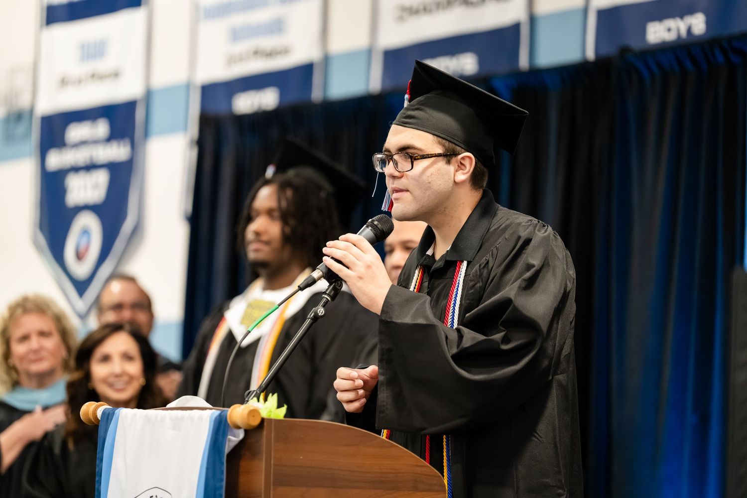 Graduate in cap and gown speaks at podium during graduation ceremony in school gymnasium.