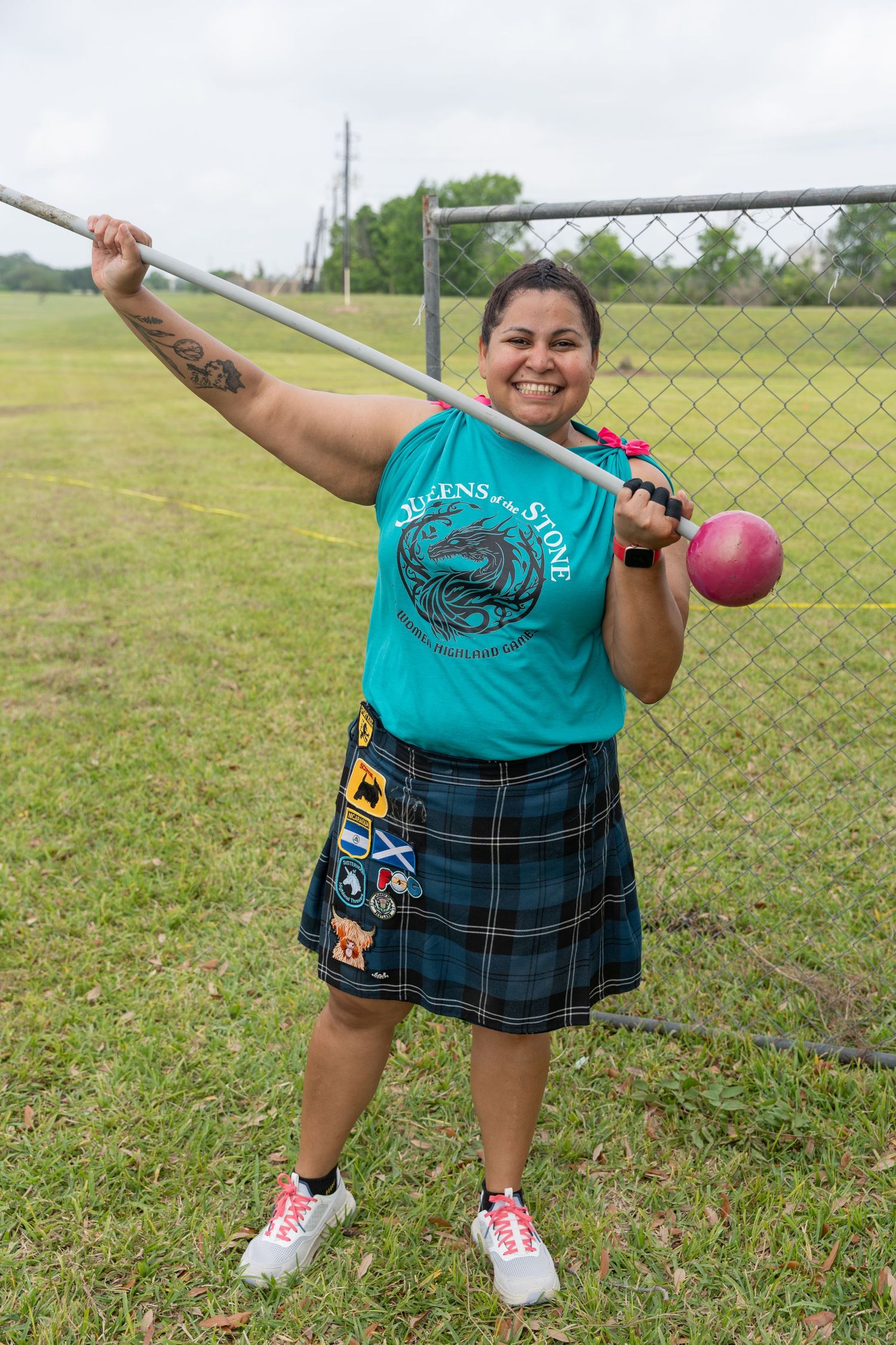 Woman in teal tank top and tartan kilt poses with hammer throw at Highland Games competition outdoors.