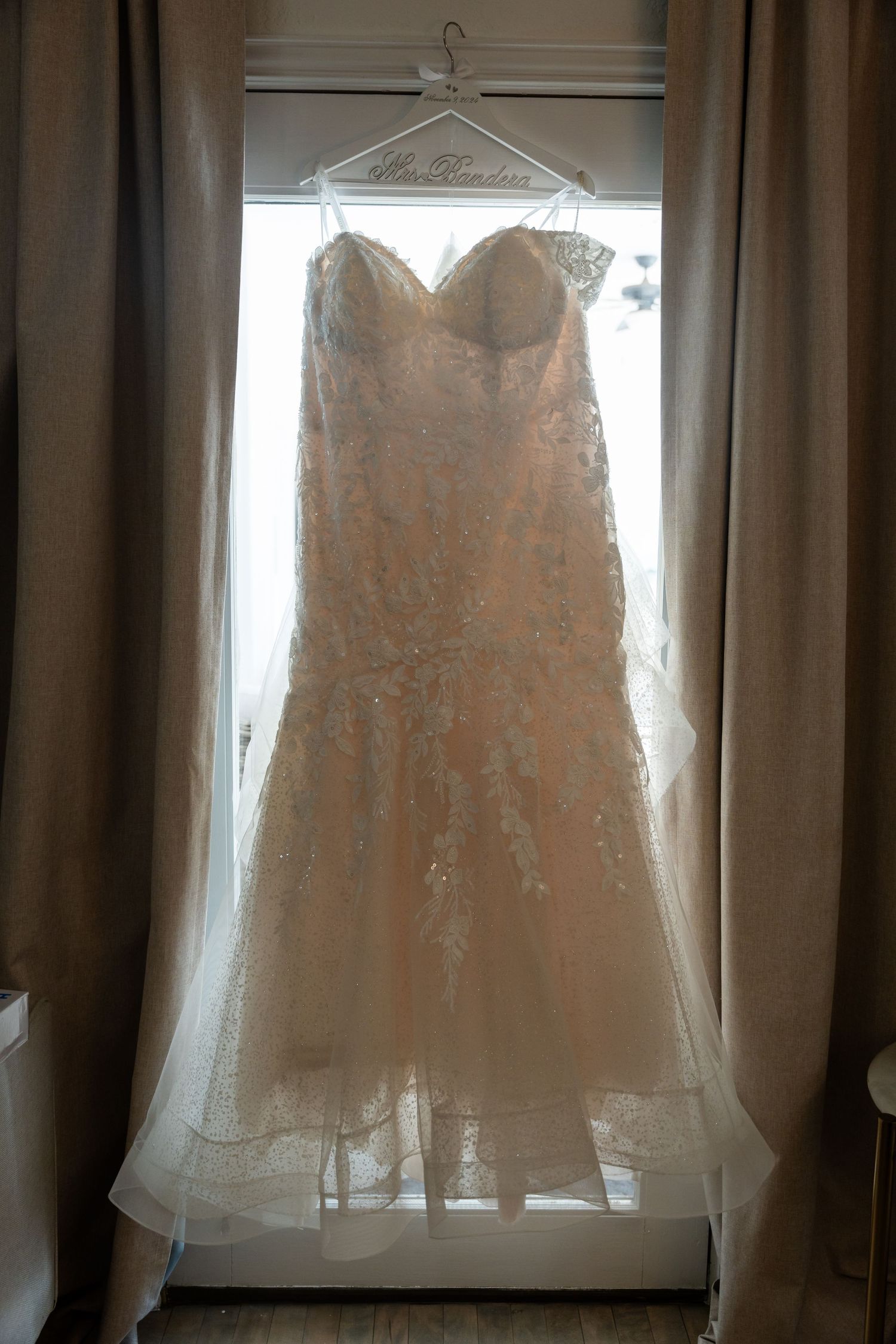 A delicate white lace wedding dress hangs in front of a window creating a romantic silhouette.