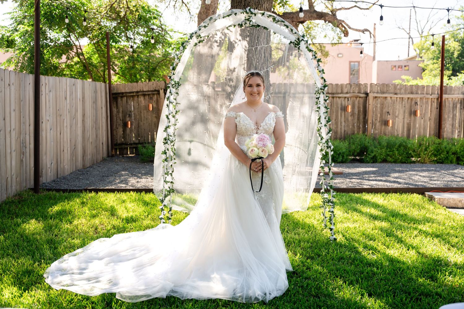 A bride stands under a floral arch in a backyard garden setting with string lights.