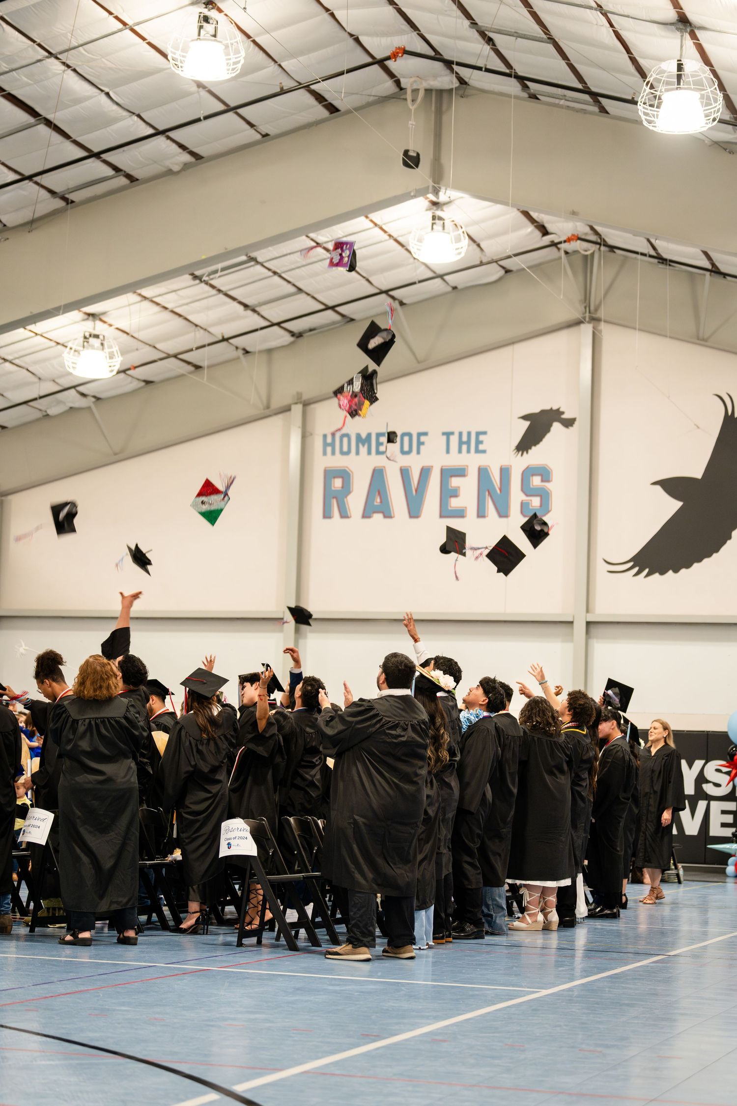 Graduates throw caps in celebration under Home of the Ravens banner in school gymnasium.