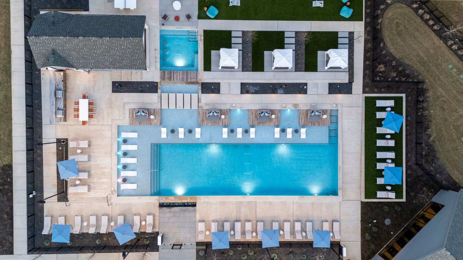 Aerial view of a modern apartment complex swimming pool with lounge chairs and cabanas arranged symmetrically around turquoise water.