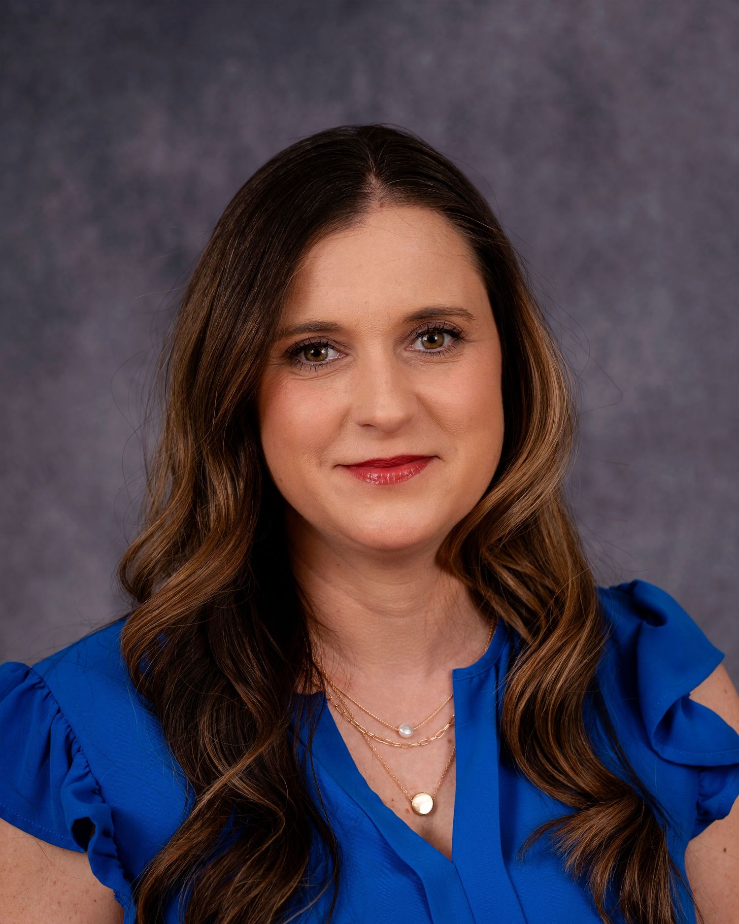 Professional portrait of person in royal blue top against mottled gray backdrop wearing delicate necklaces and styled wavy hair.