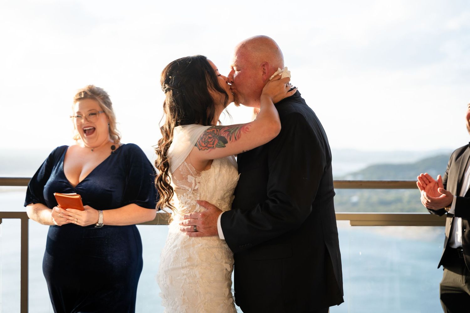 Romantic wedding kiss at scenic outdoor venue with water view.