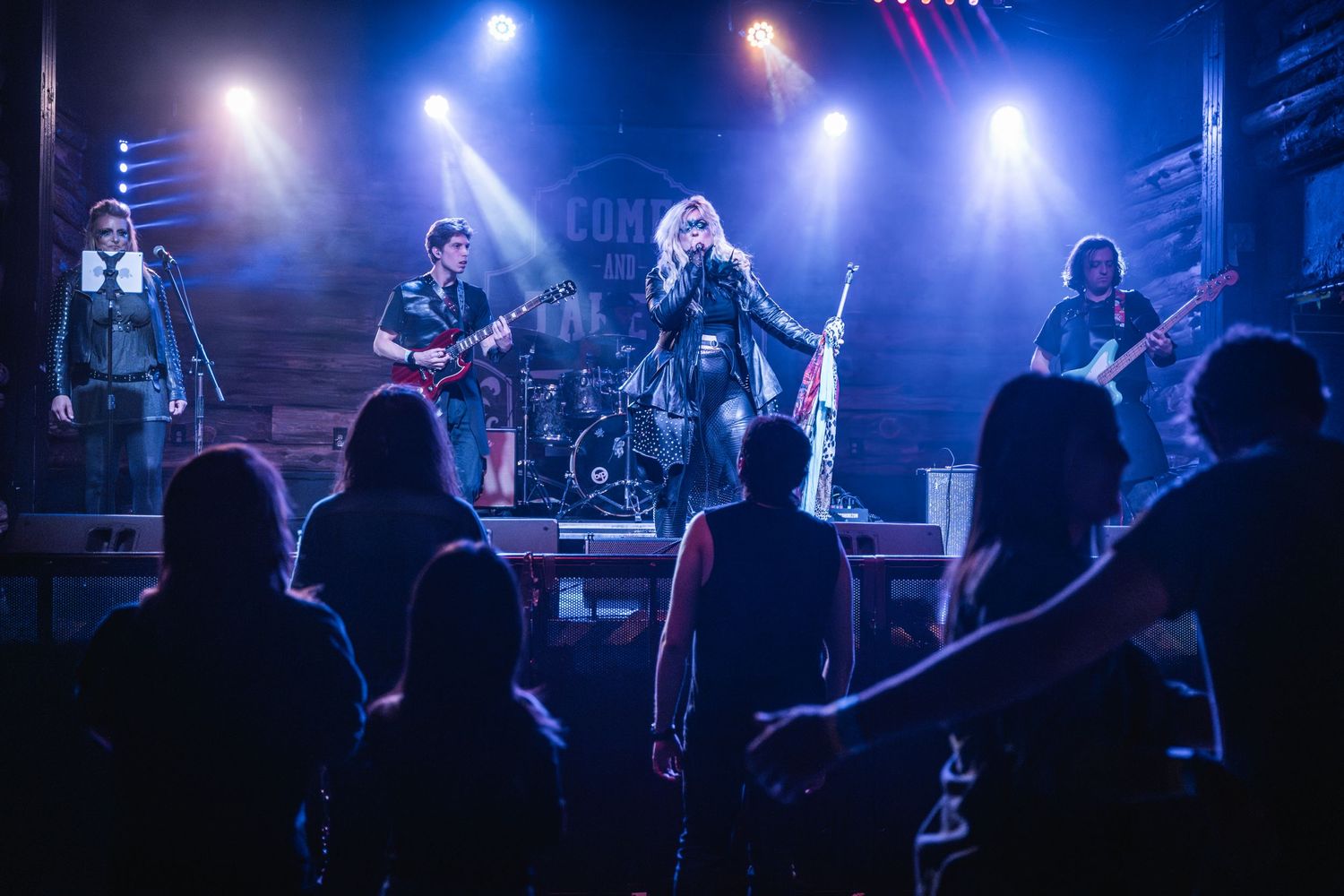 Rock band performs on stage with dramatic blue lighting and silhouetted audience members in the foreground.