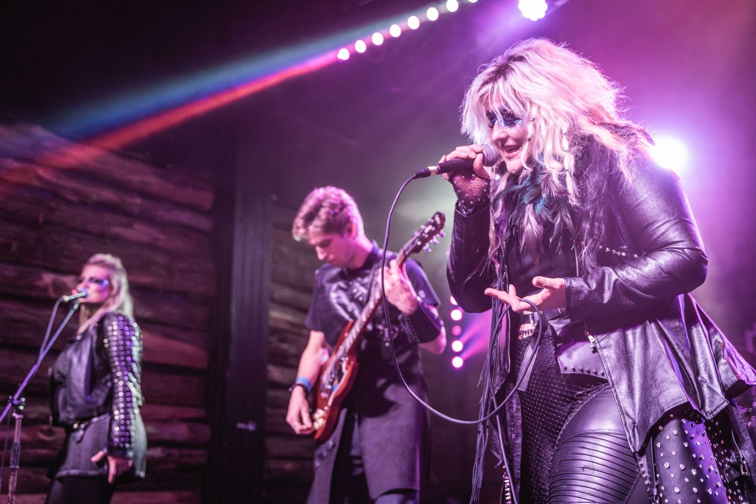 Band plays live show on wooden-walled stage with dramatic purple lighting.