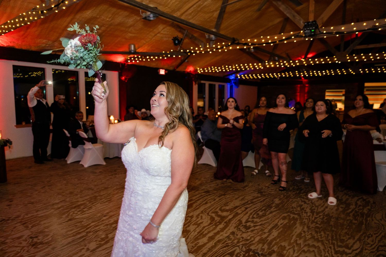A bride in a white wedding dress stands on the dance floor under string lights during the reception.