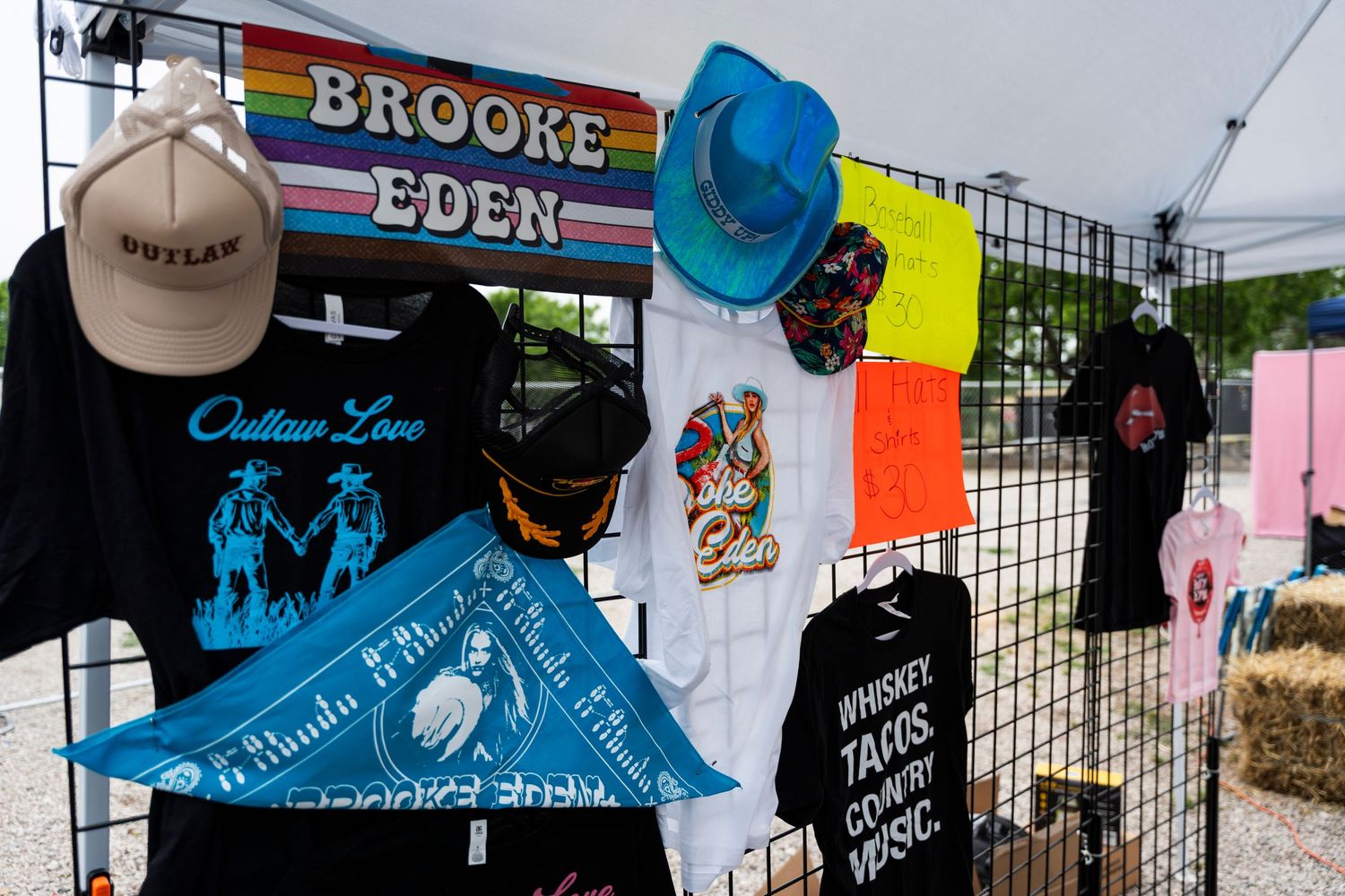 Colorful display of t-shirts and hats at a vendor booth with rainbow striped signage and vibrant merchandise.