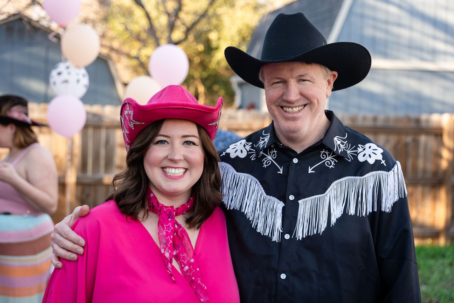 A couple in western cowboy attire smiling together at an outdoor event with pink balloons.