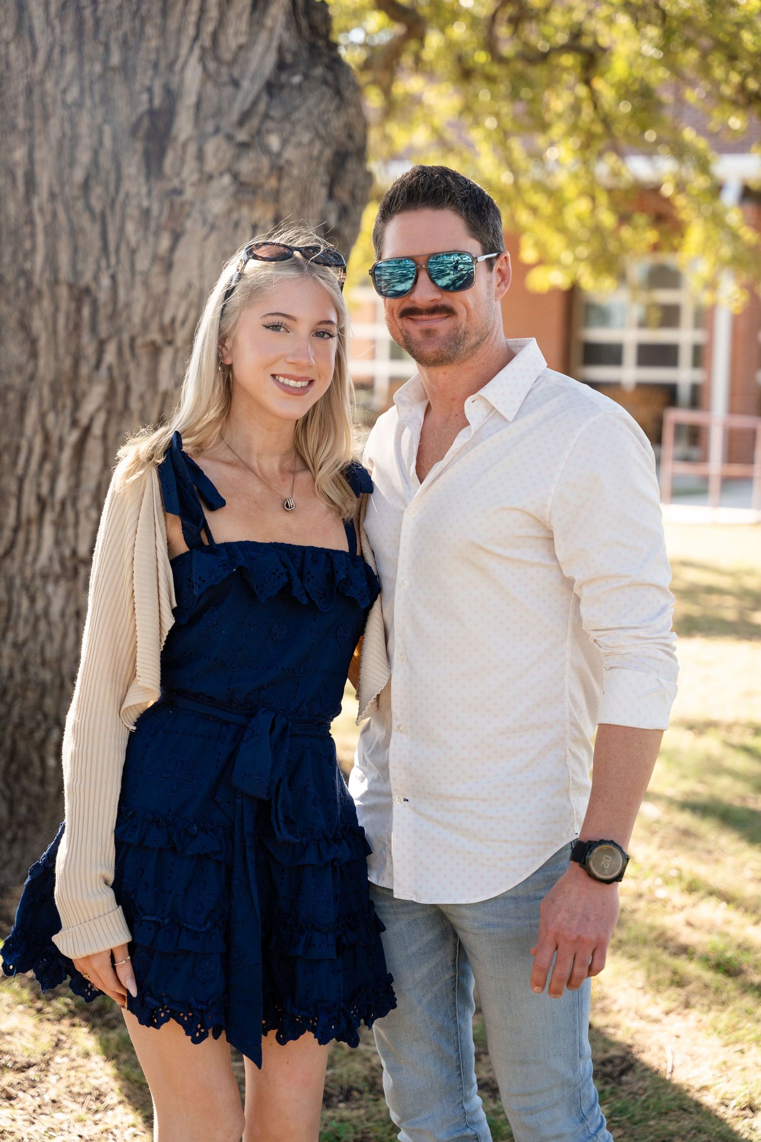 A couple poses outdoors by a tree wearing stylish casual clothing.