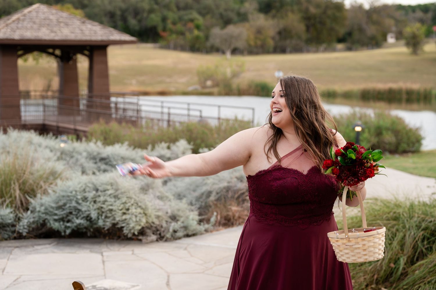 Person in burgundy dress tosses flower petals near outdoor gazebo at wedding venue.