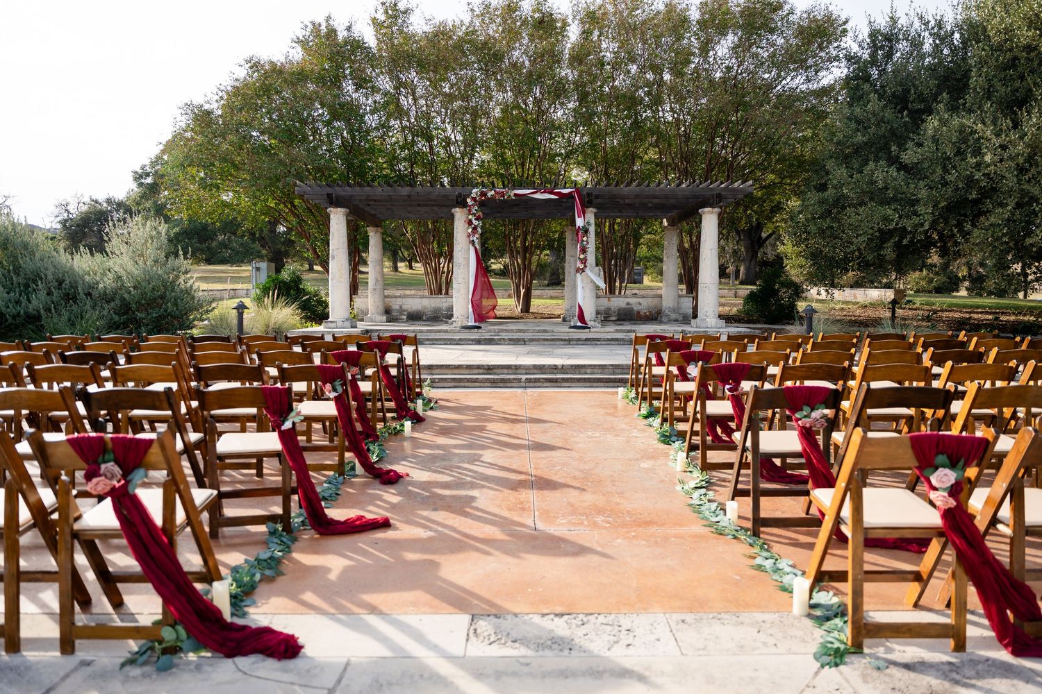 Outdoor wedding ceremony setup with burgundy-decorated wooden chairs and stone pillars.